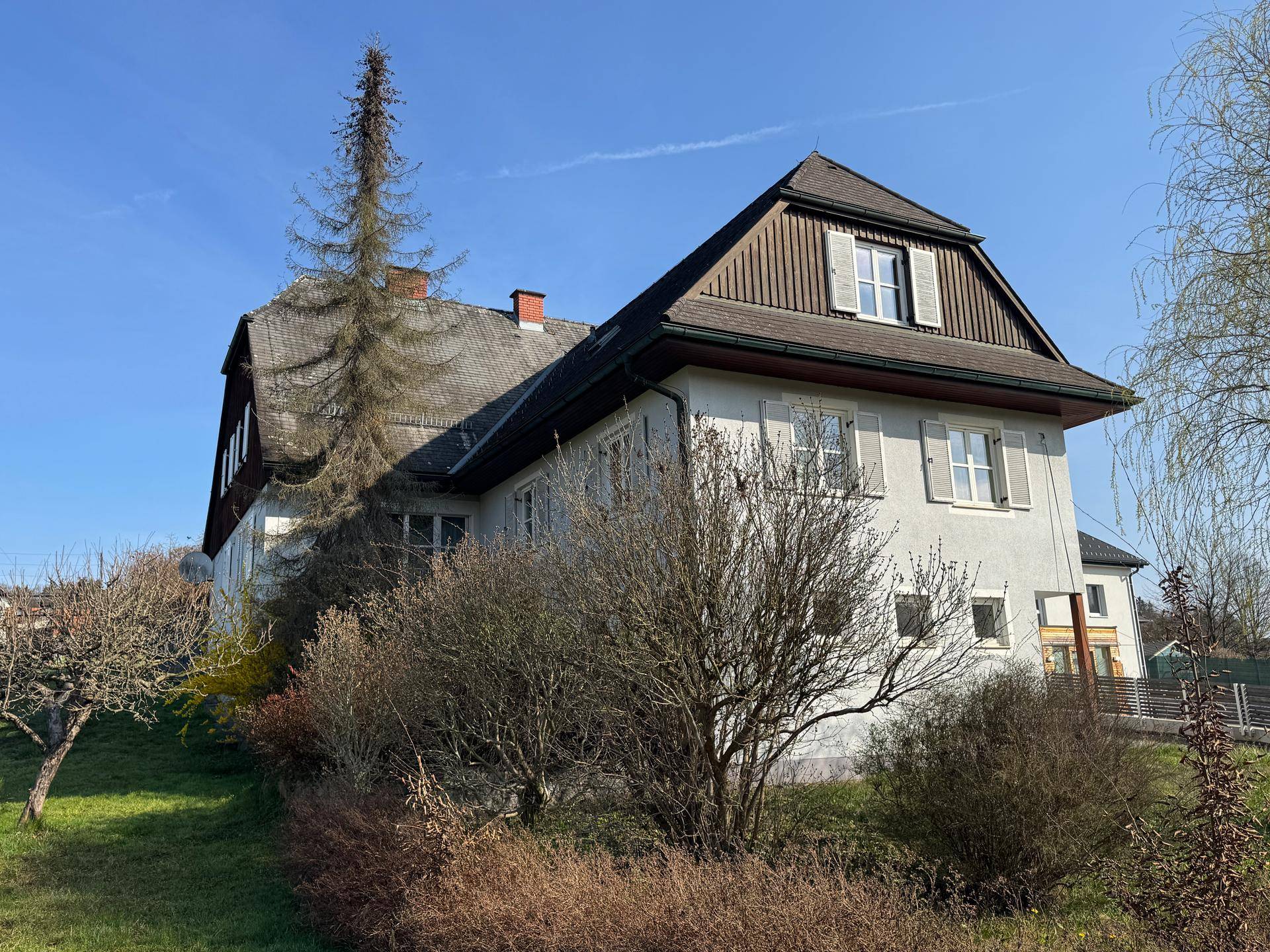 Traditionelles Haus mit Mansarddach und großem Gartenbereich unter blauem Himmel.