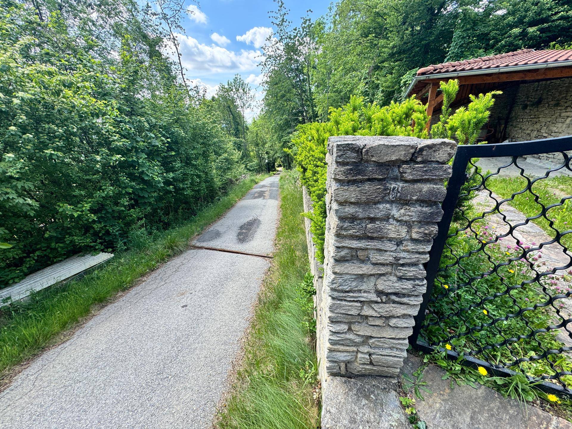 Schmaler Weg neben einer Steinmauer und einem verzierten Tor, umgeben von dichtem Grün.