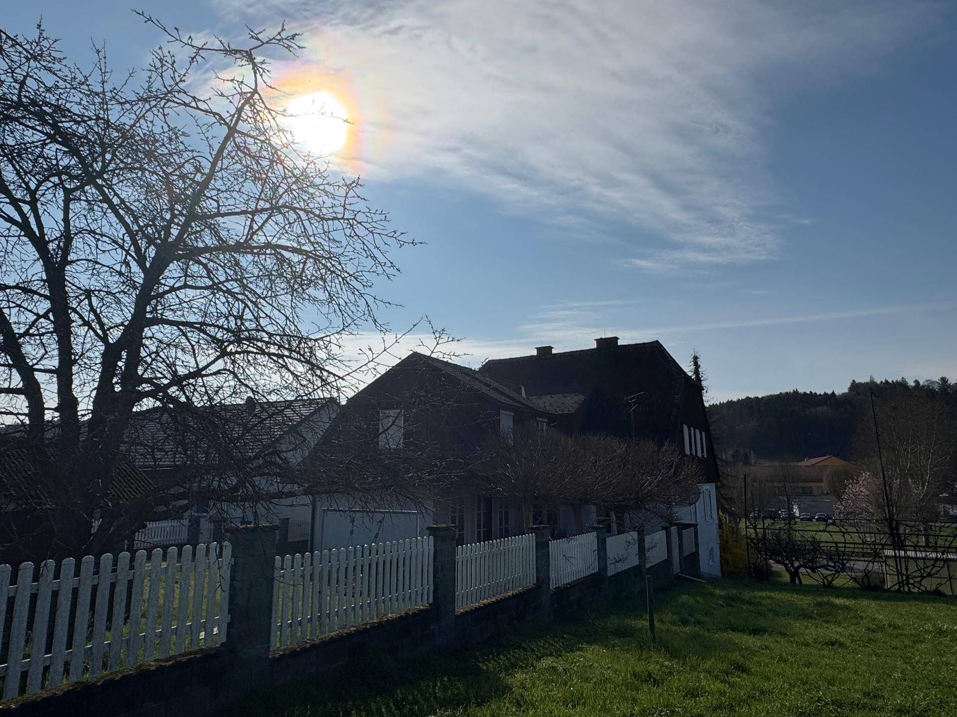 Hausansicht von hinten mit weißem Zaun und Bäumen im Gegenlicht der Sonne.