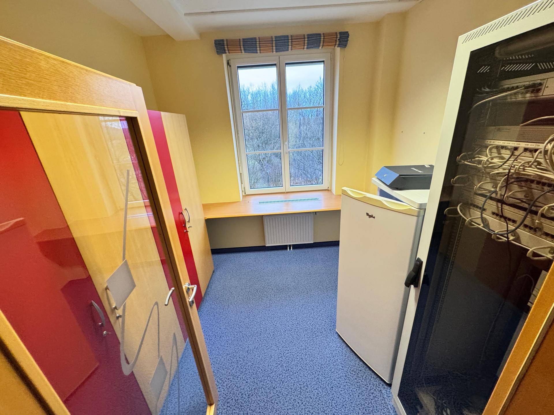 Kleiner Büroraum mit Fenster, blauem Teppichboden und einem Kühlschrank.