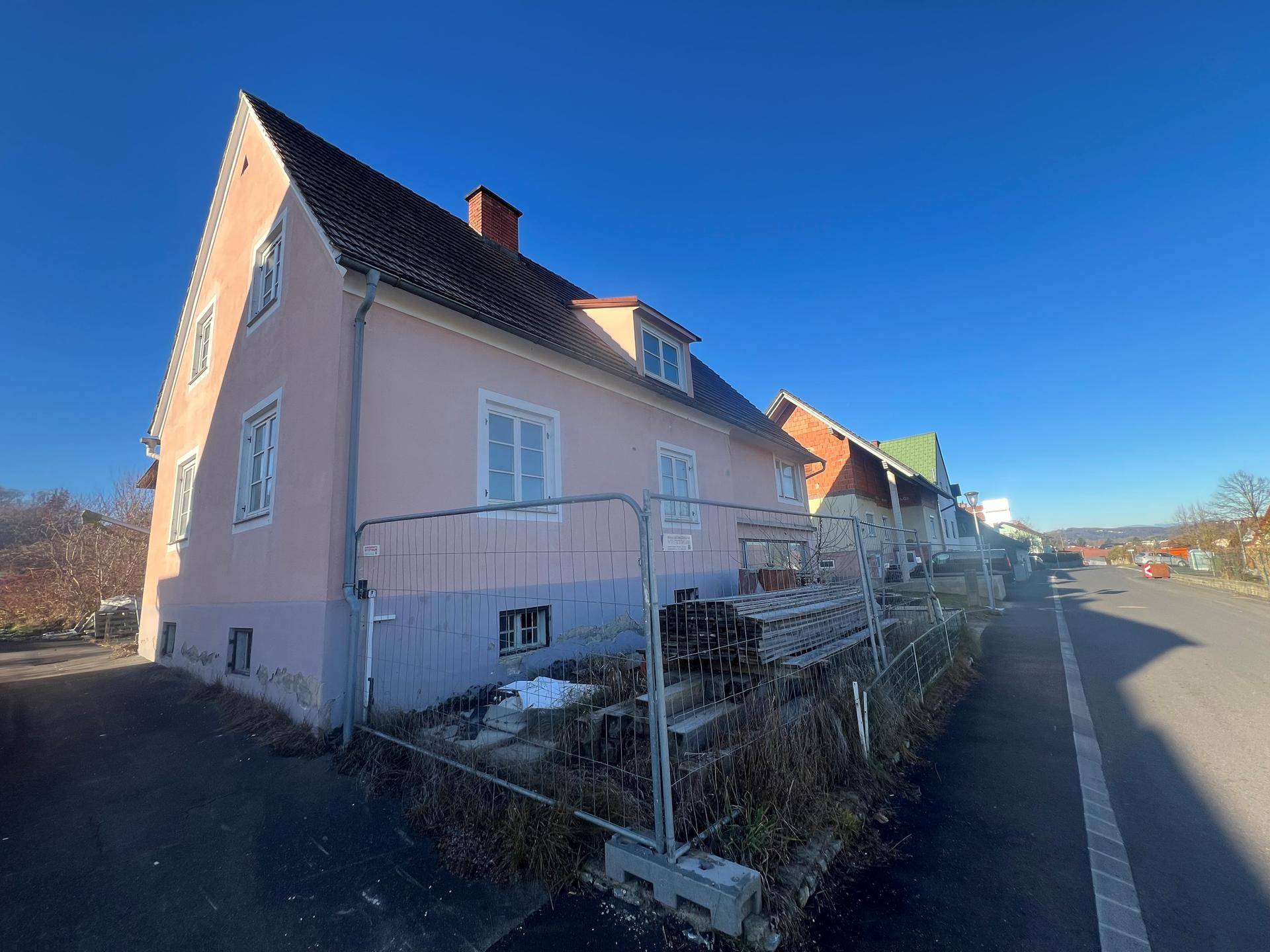 Ansicht des rosa Hauses von der Straße aus, mit Bauzaun und sonnigem Wetter.