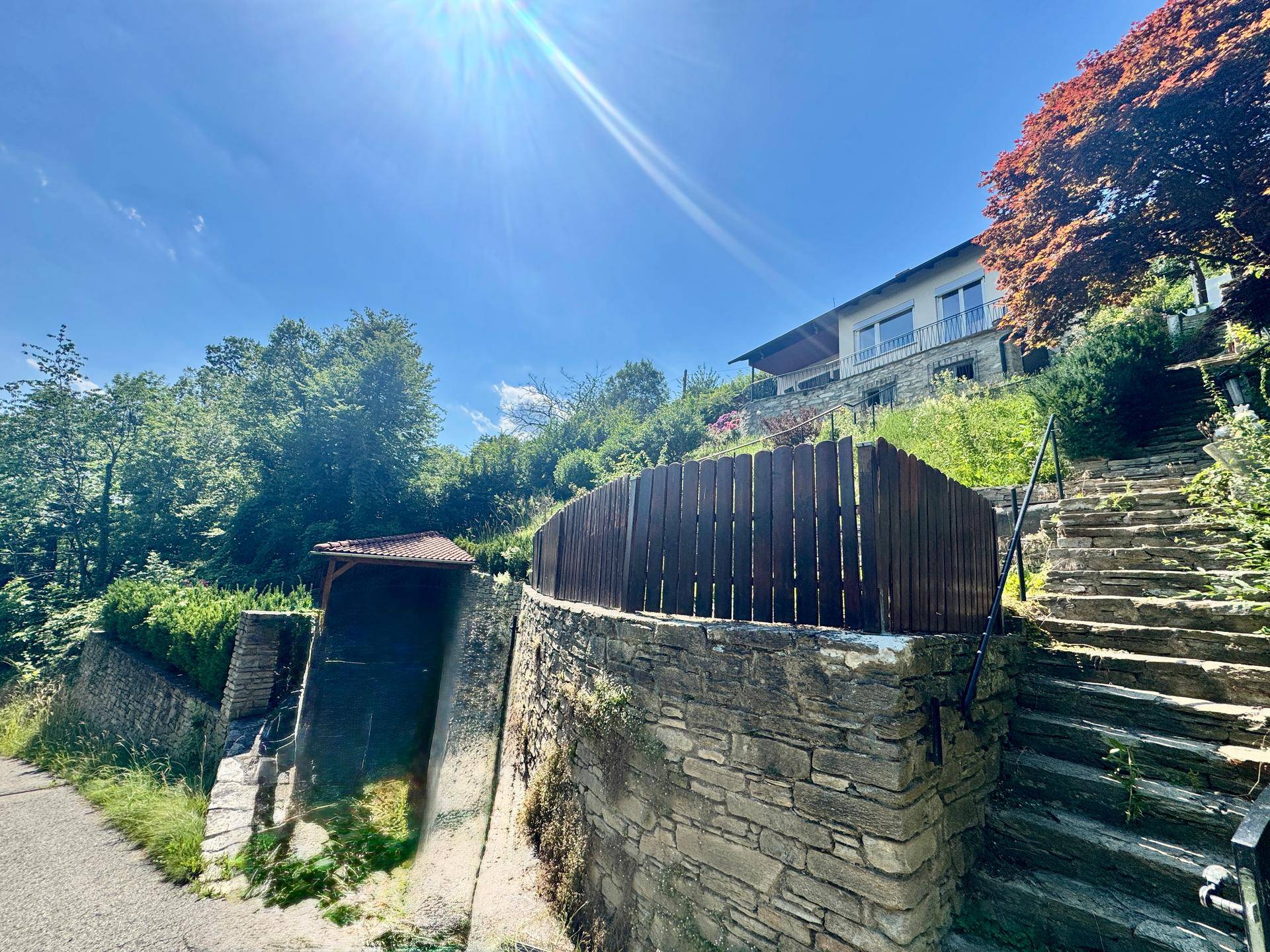 Steintreppe und Mauerwerk führen zu einem Haus auf einem bewachsenen Hang unter blauem Himmel.