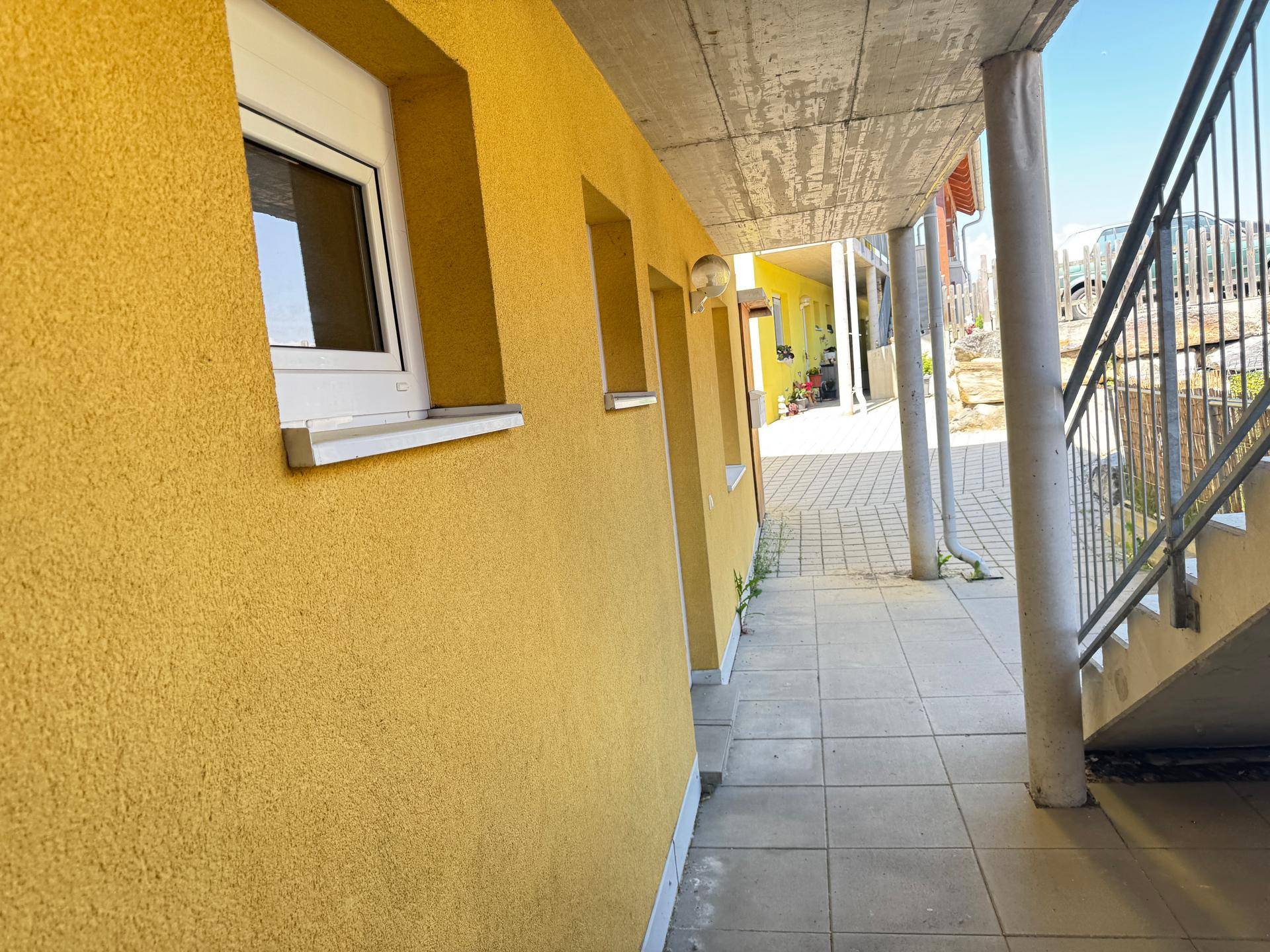 Gelbe Fassade eines Gebäudes mit weißen Fenstern und einem überdachten Außenbereich mit Treppe.