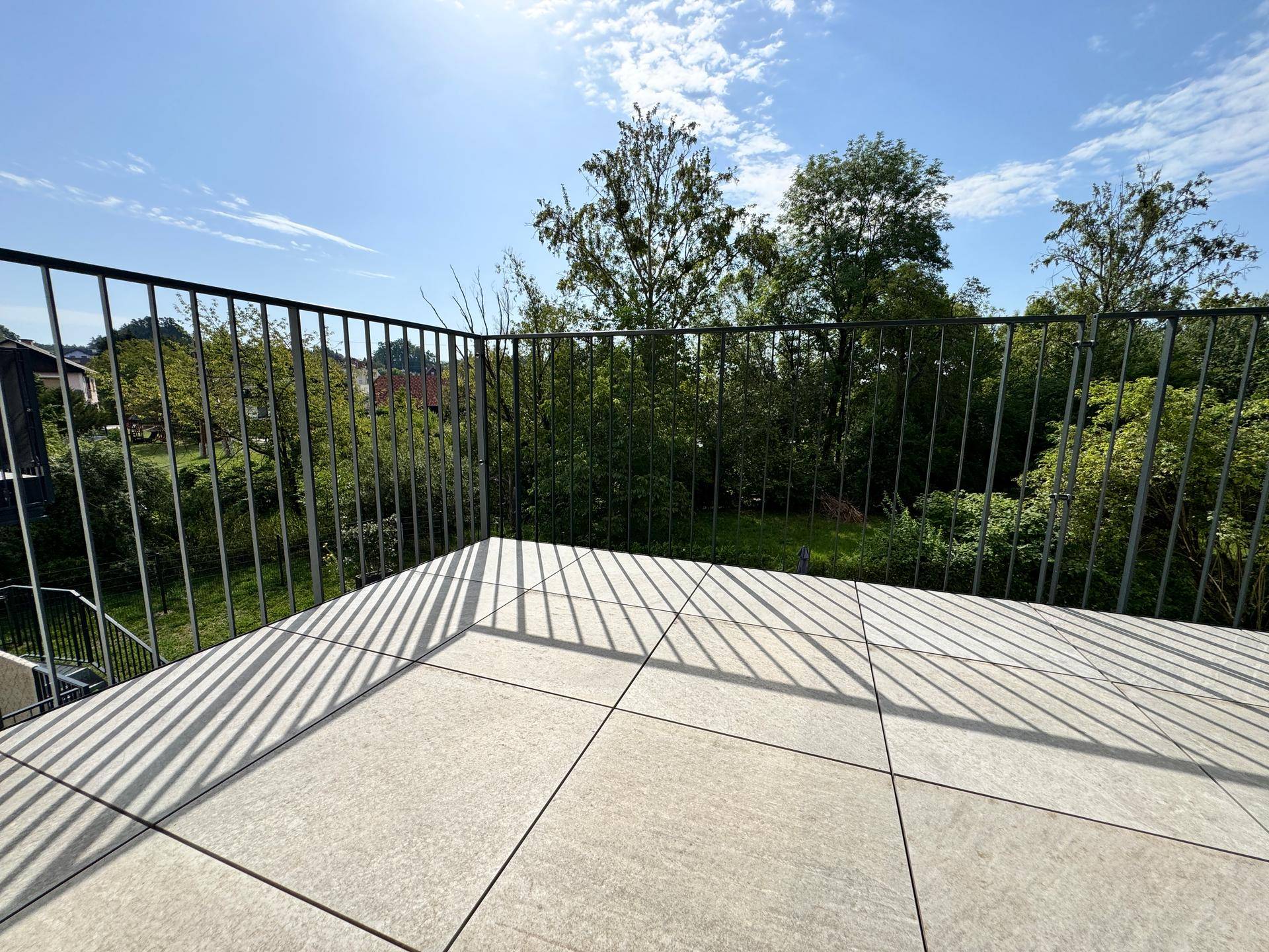 Heller Balkon mit Fliesenboden und weitem Blick ins Grüne unter blauem Himmel.