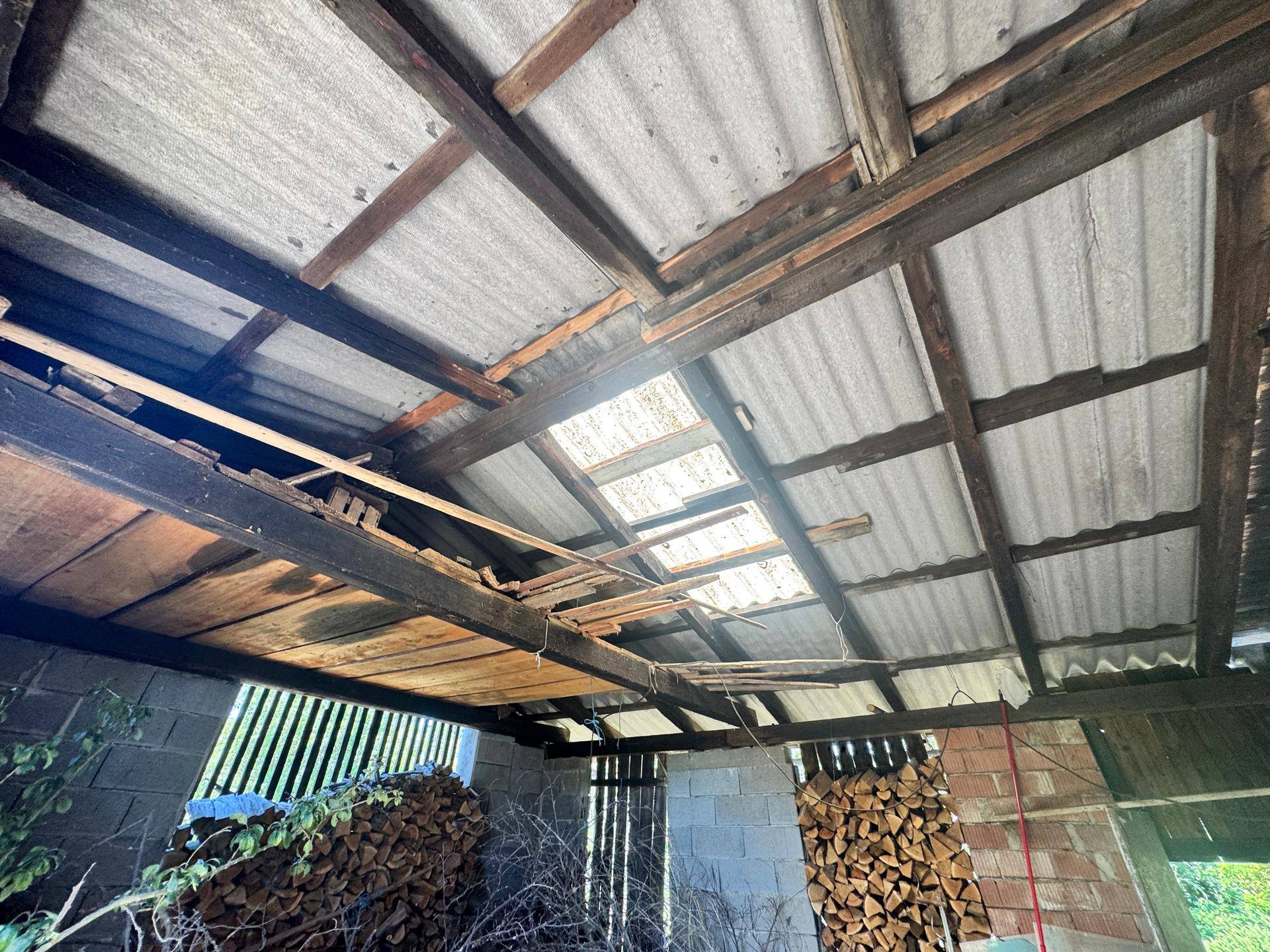 Offener Schuppen mit Holzbalken und Wellblechdach, gefüllt mit Brennholz und Ästen.