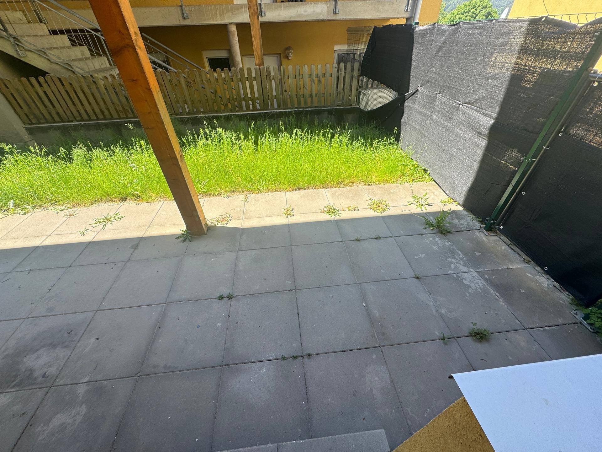 Gepflasterte Terrasse mit Rasenfläche und einem schwarzen Sichtschutzzaun im Gartenbereich.