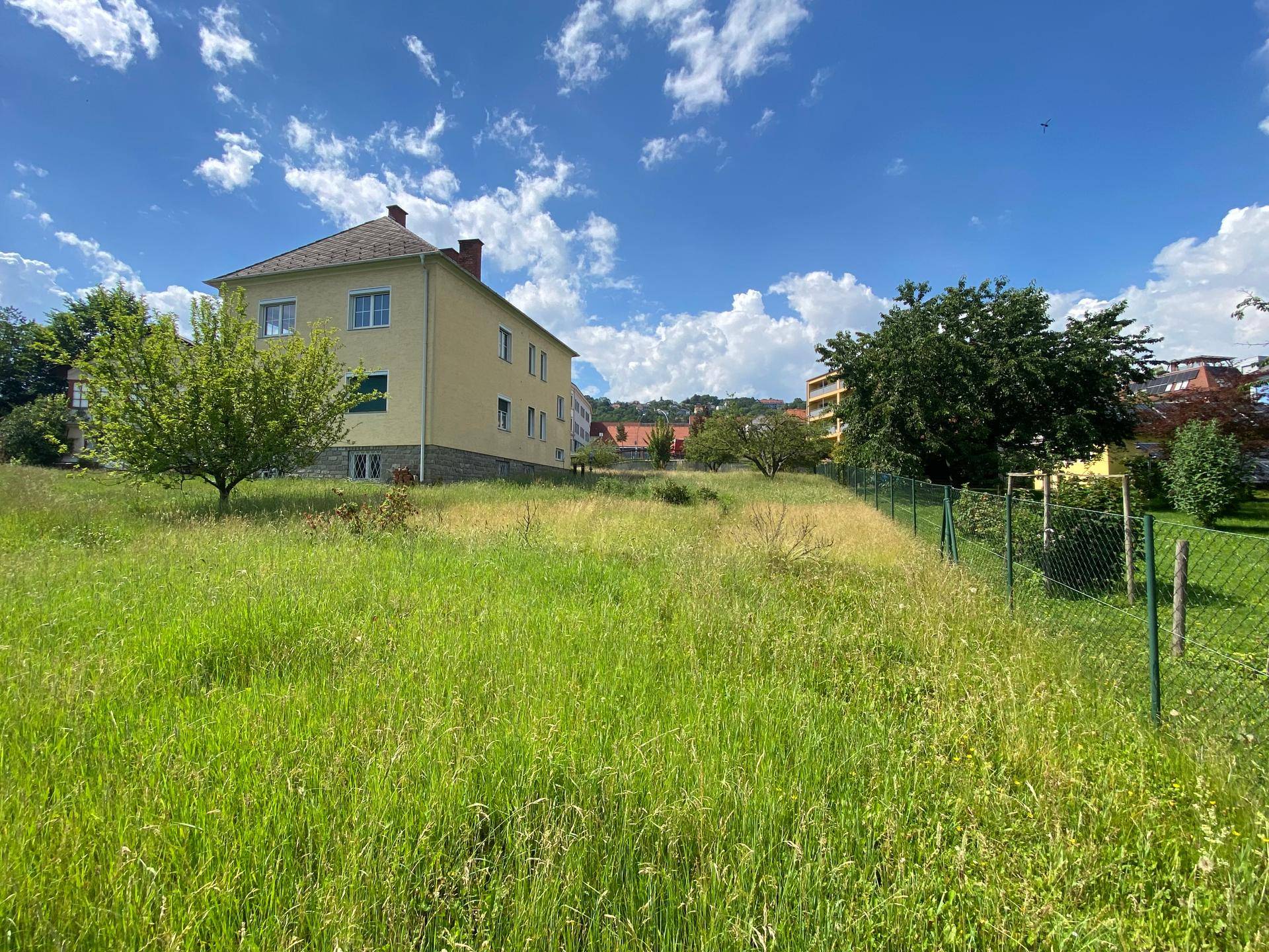 Gelbes Wohnhaus mit grüner Wiese und Zaun im Vordergrund, umgeben von weiteren Gebäuden.