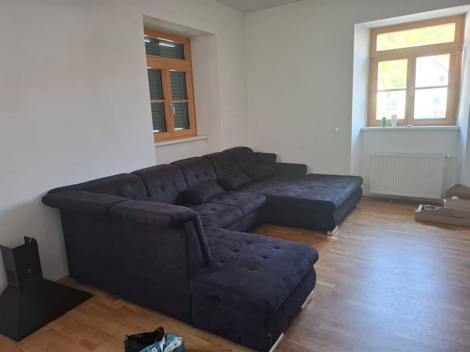 Helles Wohnzimmer mit bequemer Eckcouch, Parkettboden und Heizkörper unter dem Fenster.