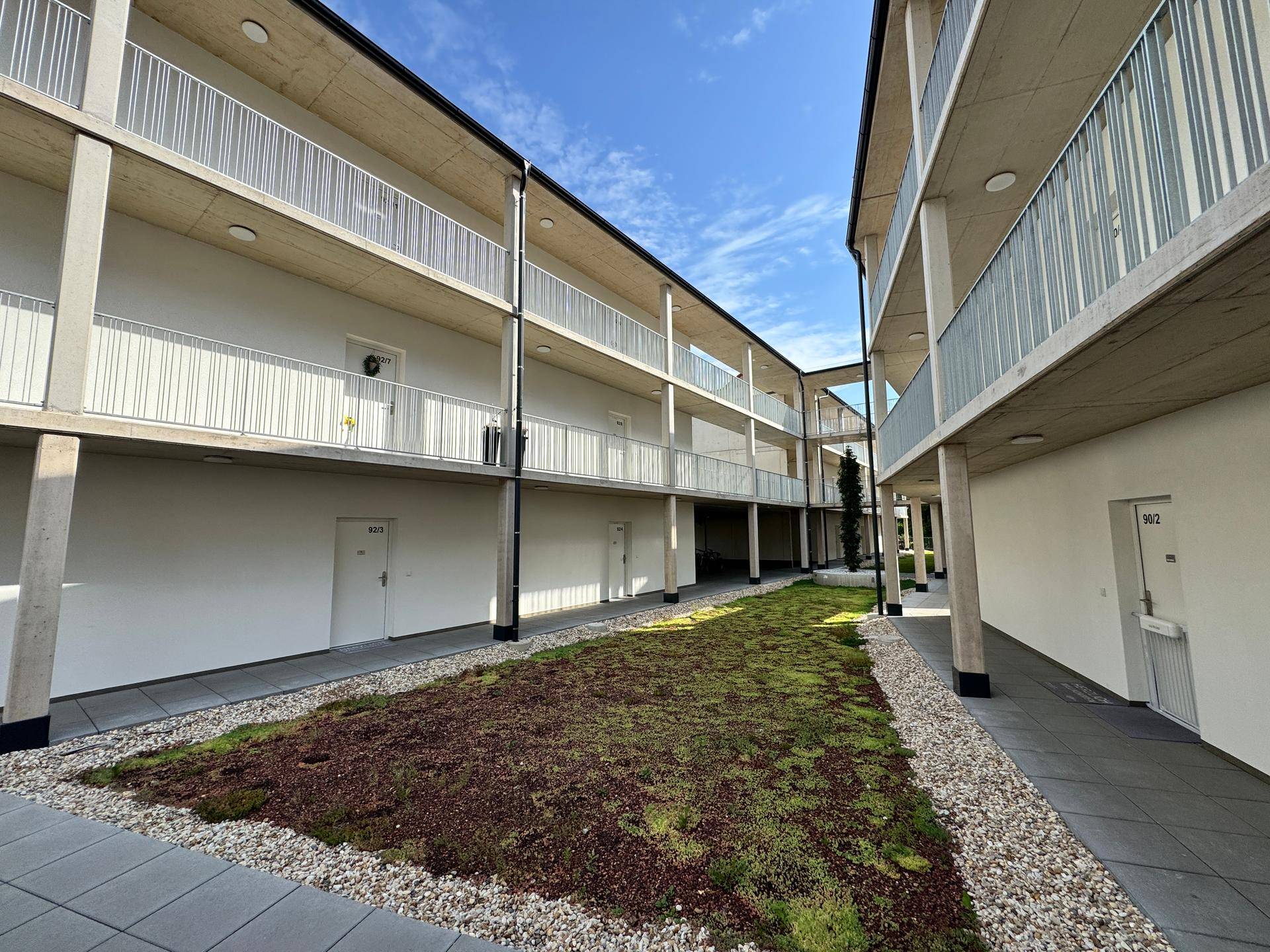 Innenhof zwischen zwei modernen Apartmentgebäuden mit begrünten Flächen und gepflasterten Gehwegen.