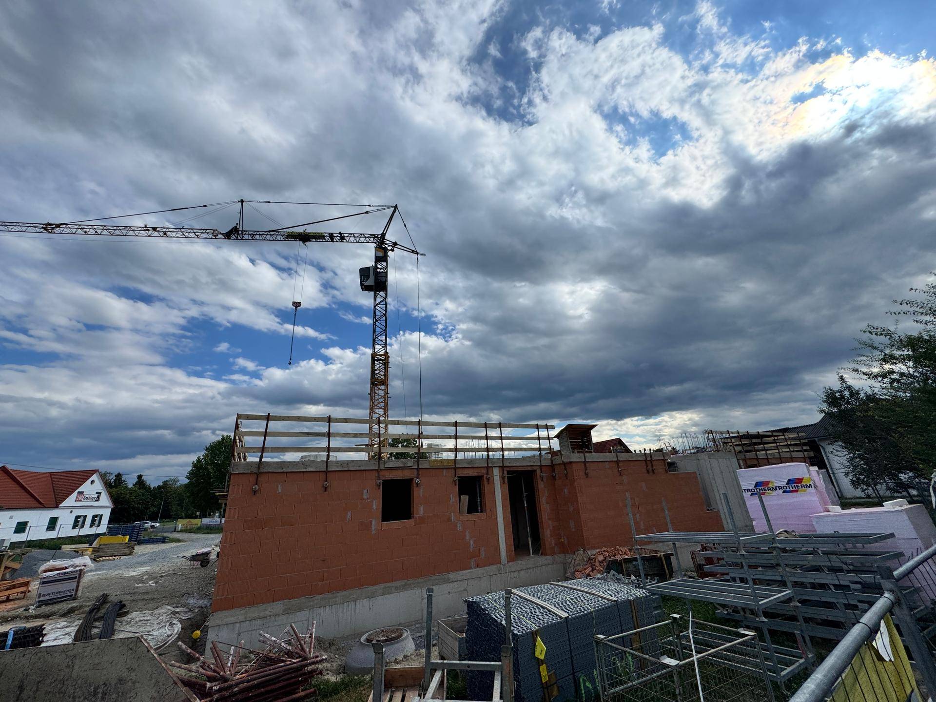 Baustelle mit Rohbau, Kran und Baumaterialien unter einem dramatischen, bewölkten Himmel.