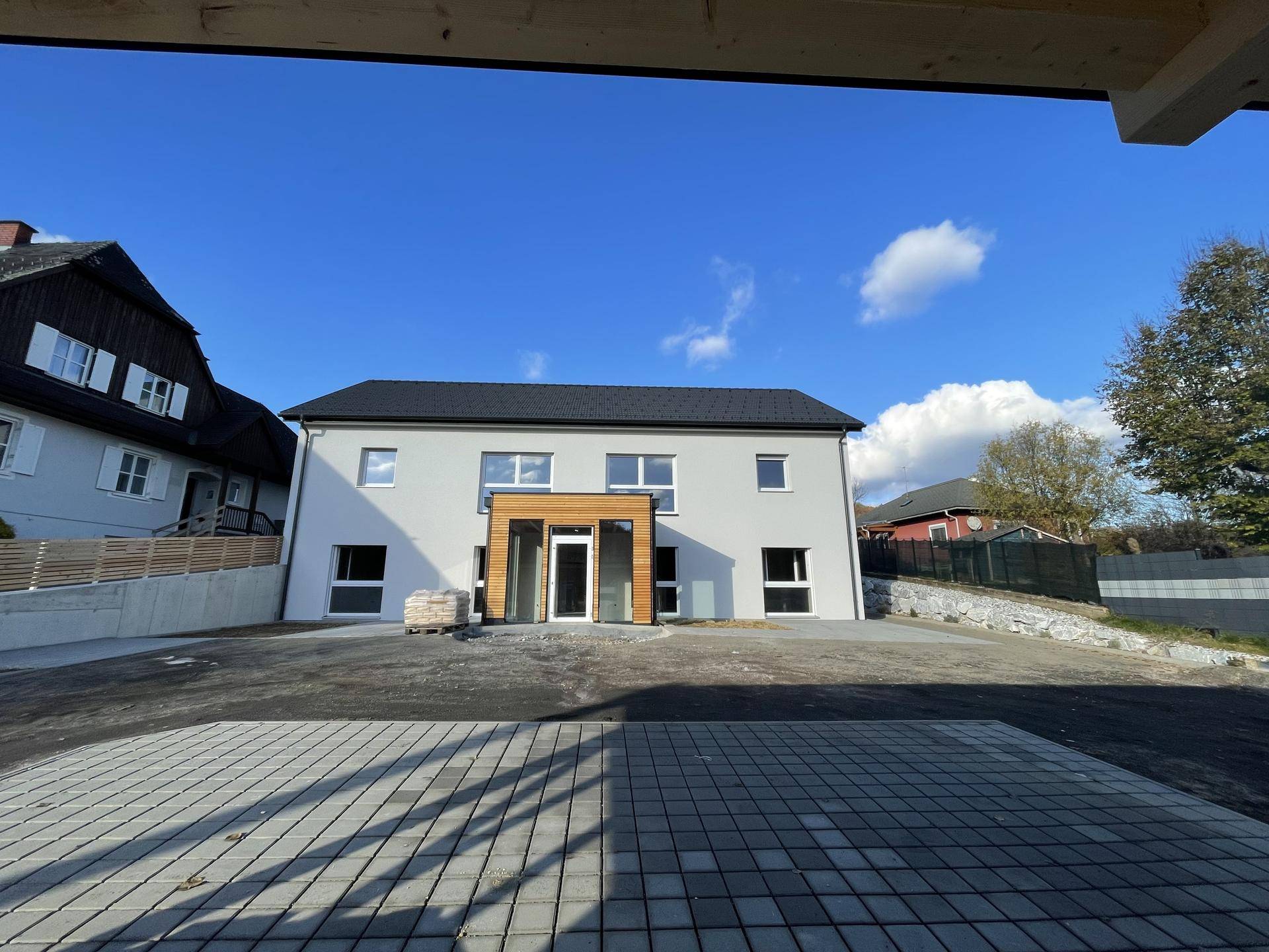 Moderner Neubau mit gepflastertem Vorplatz und umliegender Landschaft unter blauem Himmel.