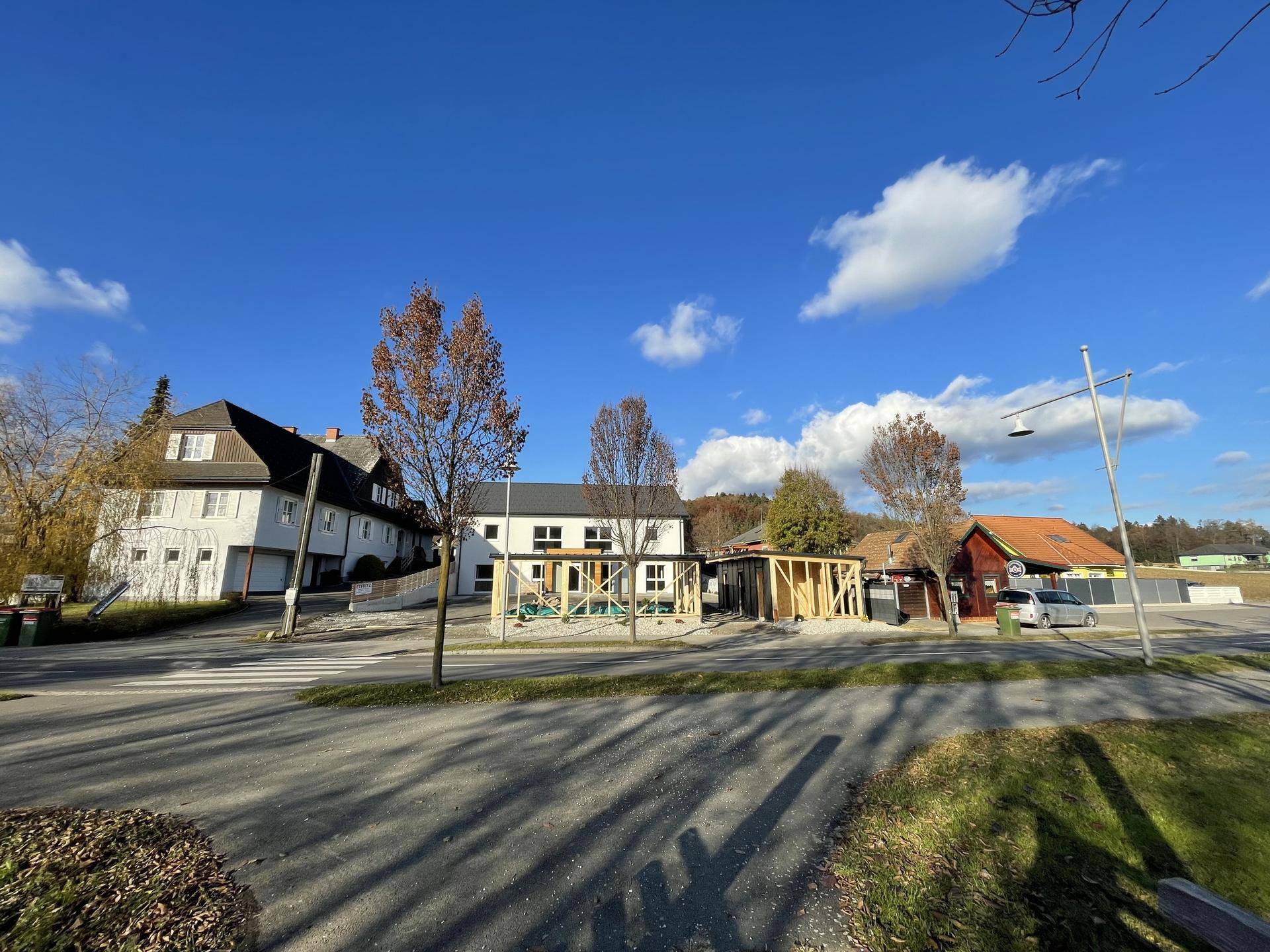 Neubau in einer ländlichen Umgebung mit weiteren Gebäuden und Bäumen unter blauem Himmel.