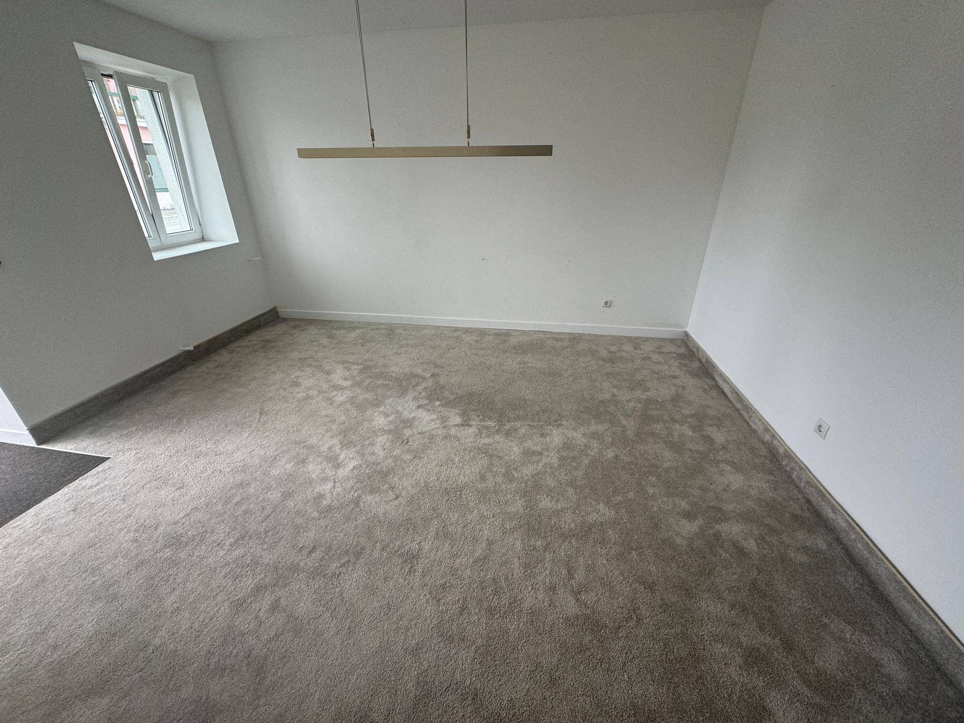 Heller, leerer Raum mit grauem Teppichboden und einem modernen Deckenlicht, bereit zur Einrichtung.