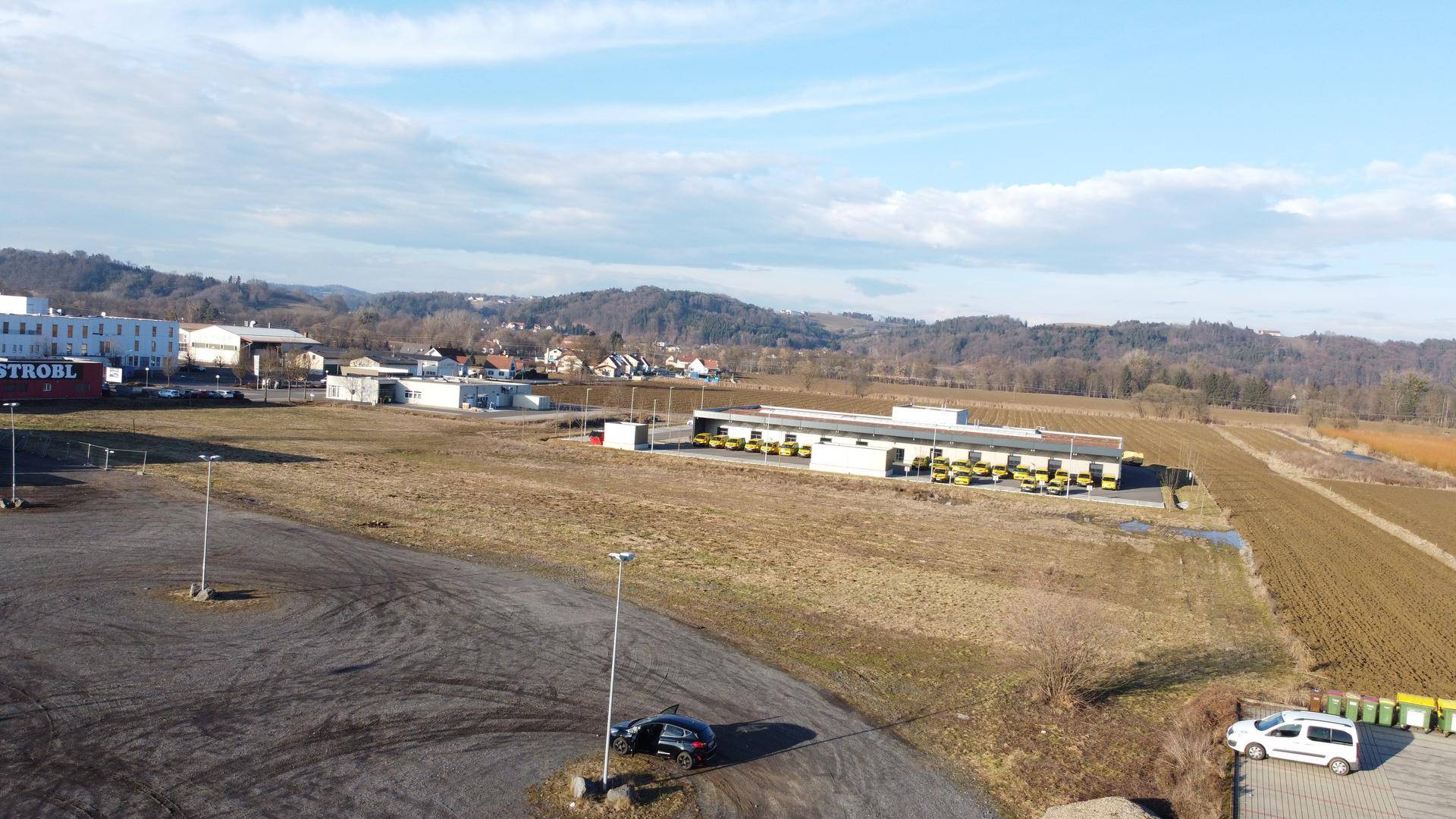 Luftaufnahme eines großen unbebauten Grundstücks mit angrenzenden Gewerbegebäuden und ländlicher Umgebung unter blauem Himmel.