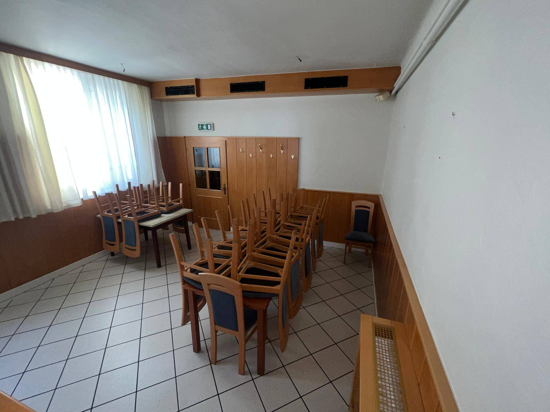 Speisesaal mit gestapelten Tischen und Stühlen, Holzvertäfelung und großen Fenstern.