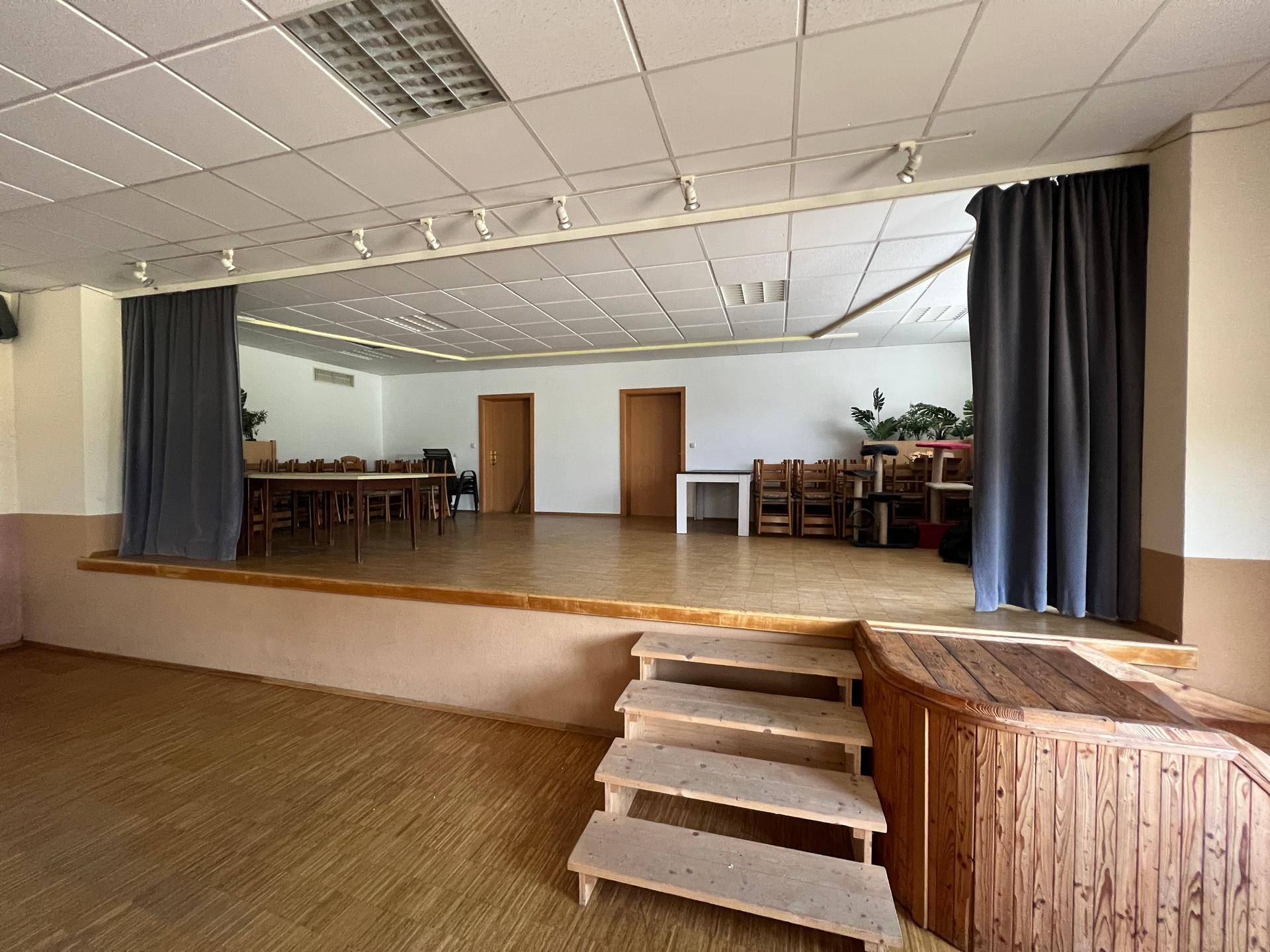 Großer Veranstaltungssaal mit Holzbühne, Vorhängen und vielen Tischen und Stühlen im Hintergrund.