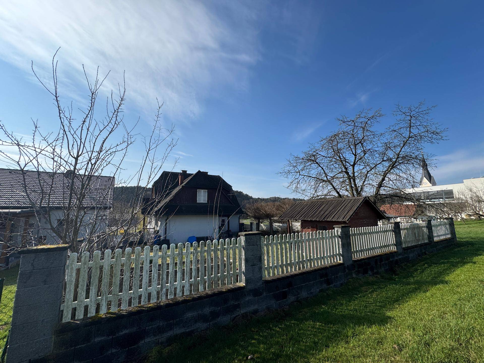 Hausansicht von der Gartenseite mit weißem Zaun und einem Kirchturm in der Ferne.