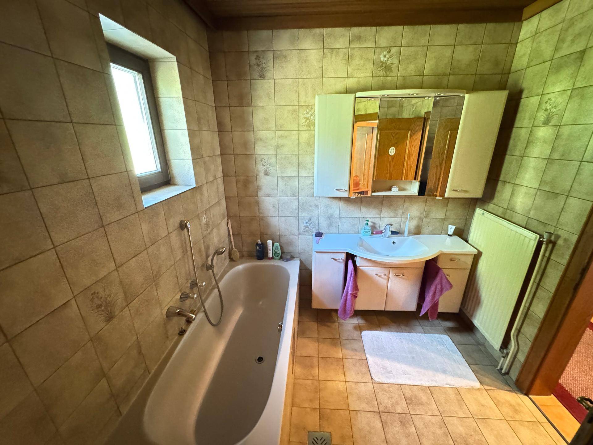 Badezimmer mit Badewanne, Waschbecken, Spiegelschrank und Fliesen an Wänden und Boden.