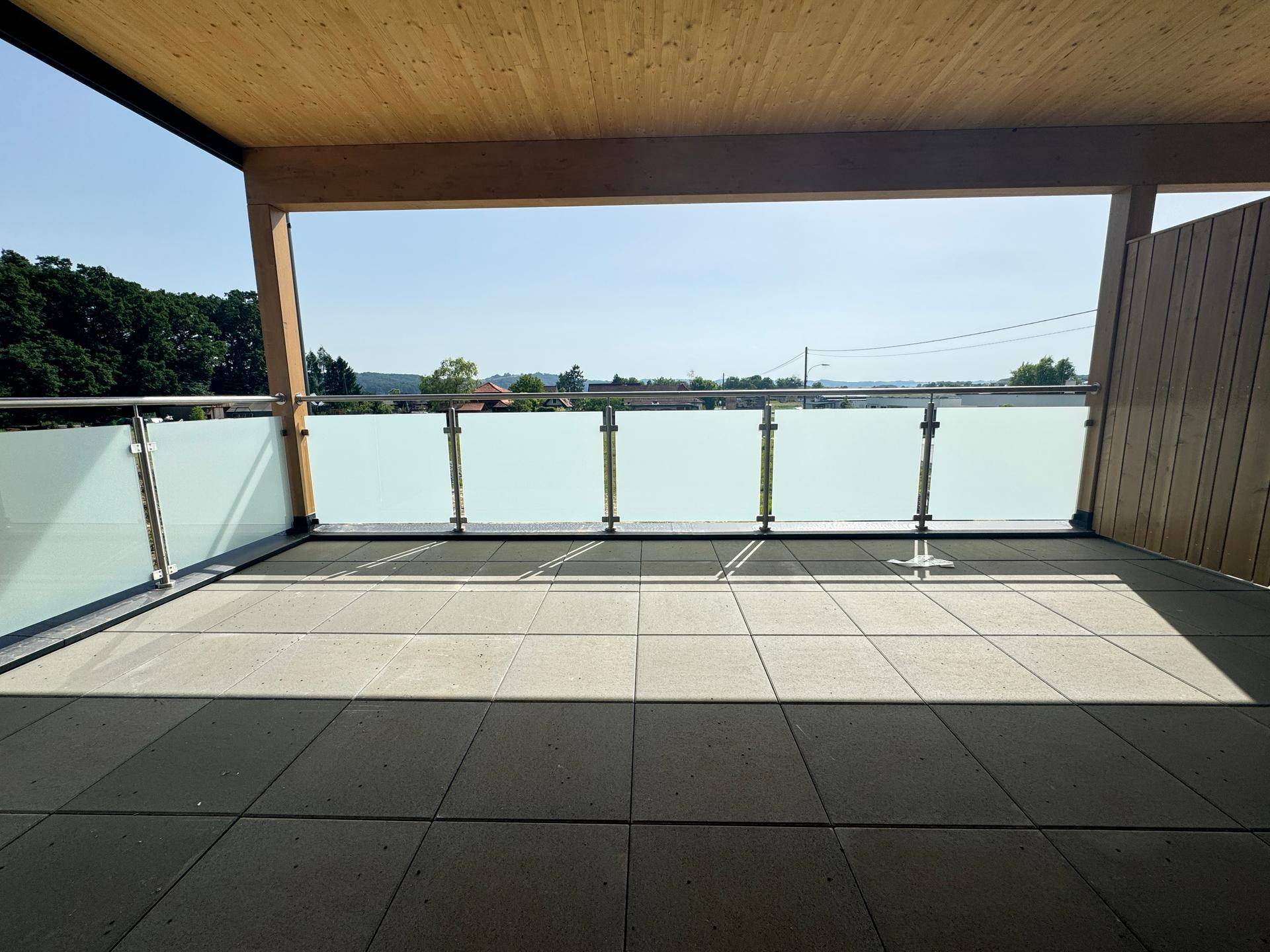 Überdachter Balkon mit Fliesenboden, Glasgeländer und sonnigem Blick in die Ferne.