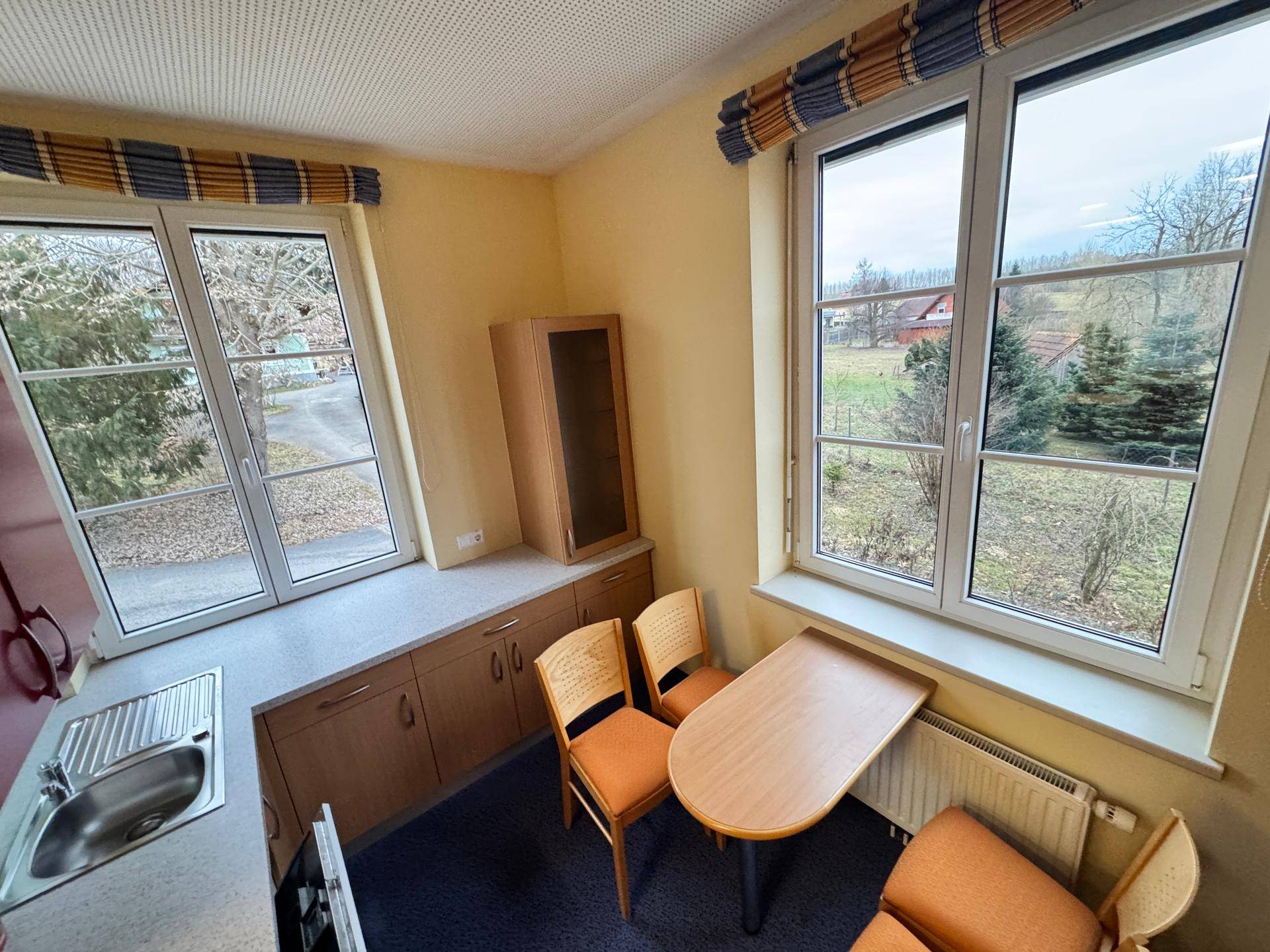 Kleine Küche mit Spüle, Unterschränken und Essbereich am Fenster mit Blick ins Grüne.