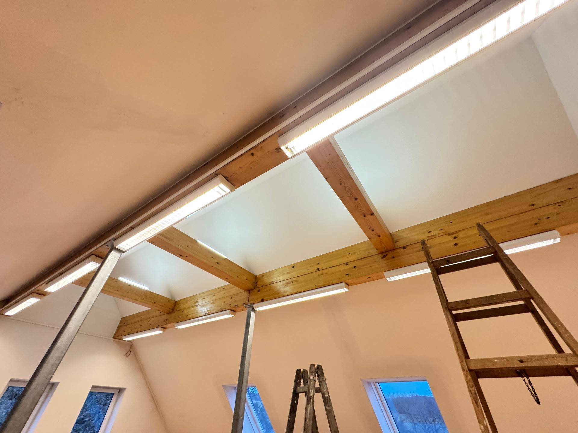 Deckenansicht eines Dachbodenraums mit sichtbaren Holzbalken und Leuchtstoffröhren.