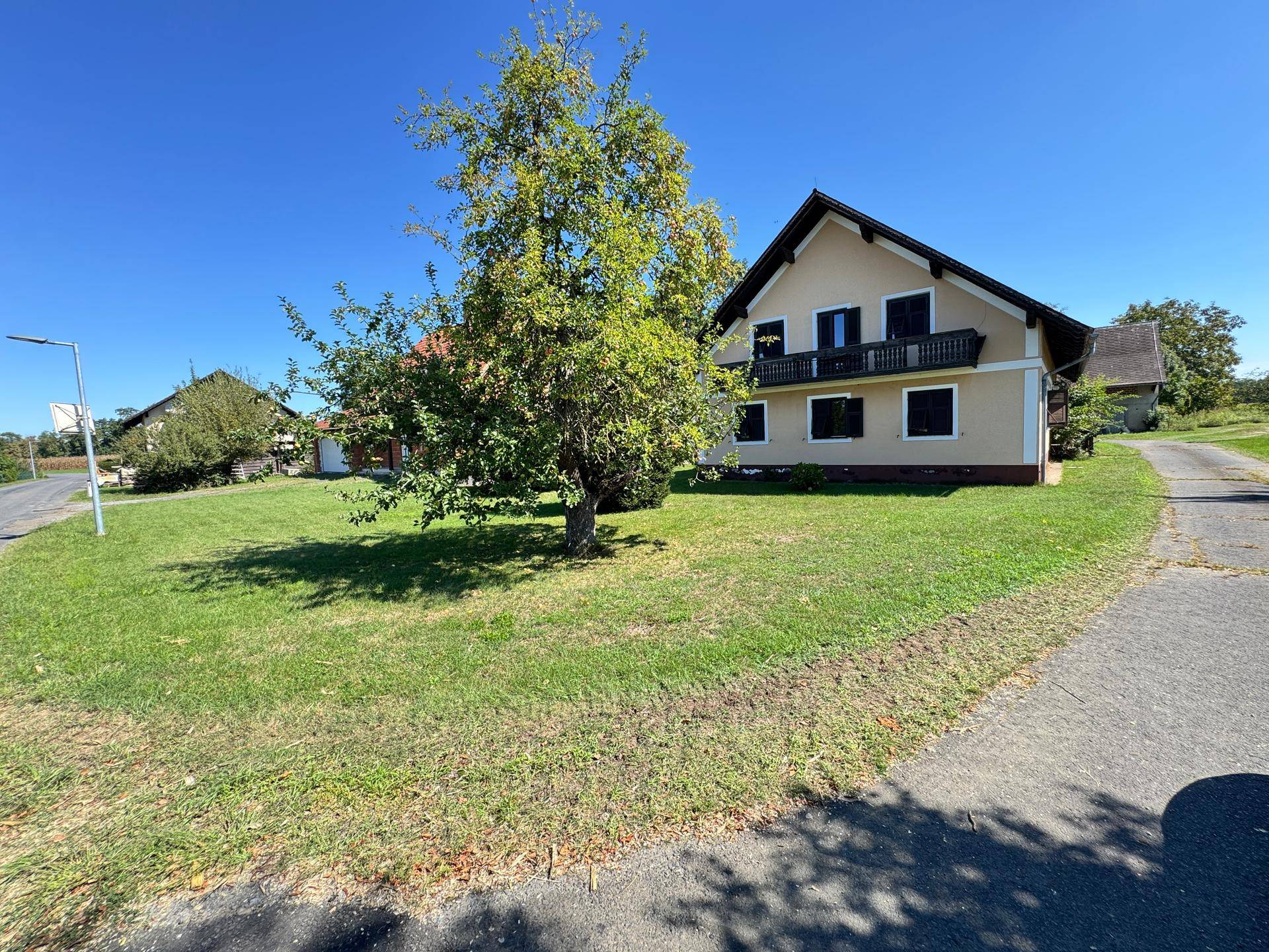 Haus mit Balkon, grüner Wiese und Zufahrtsweg in ländlicher Umgebung unter blauem Himmel.