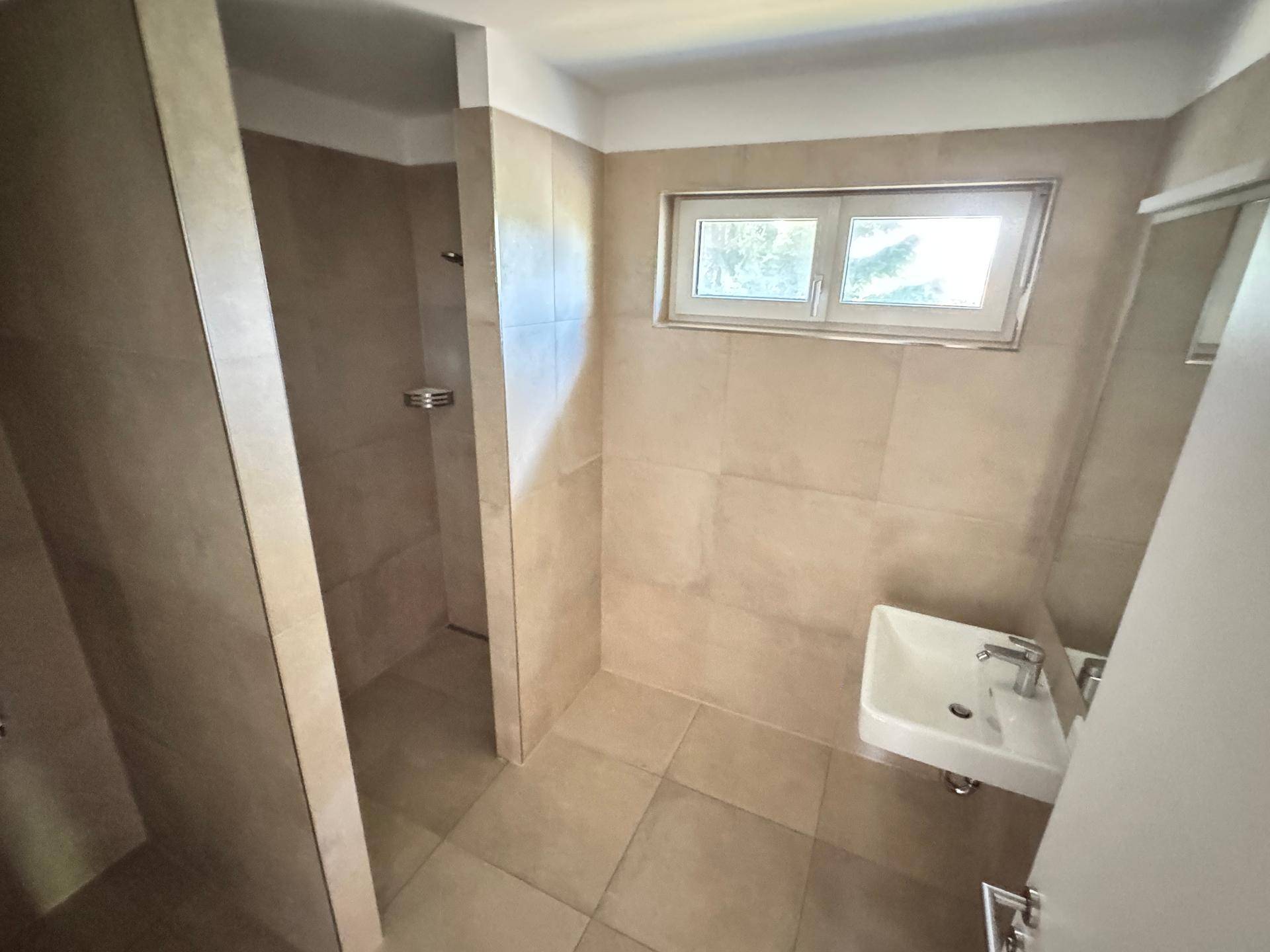 Modernes Badezimmer mit begehbarer Dusche, Waschbecken und Fenster, alles in beigefarbenen Fliesen.