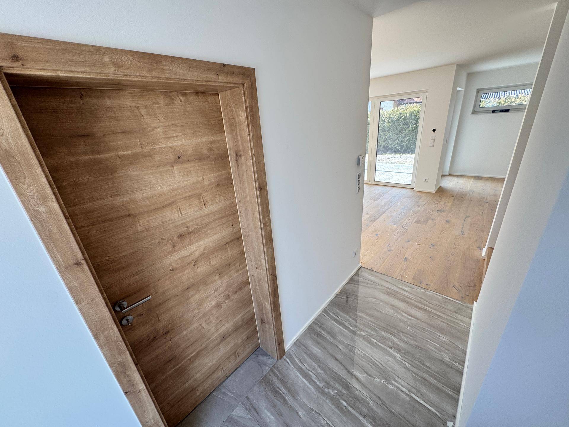 Heller Flur mit Holztür und Blick auf den Wohnbereich mit Holzboden und großen Fenstern.