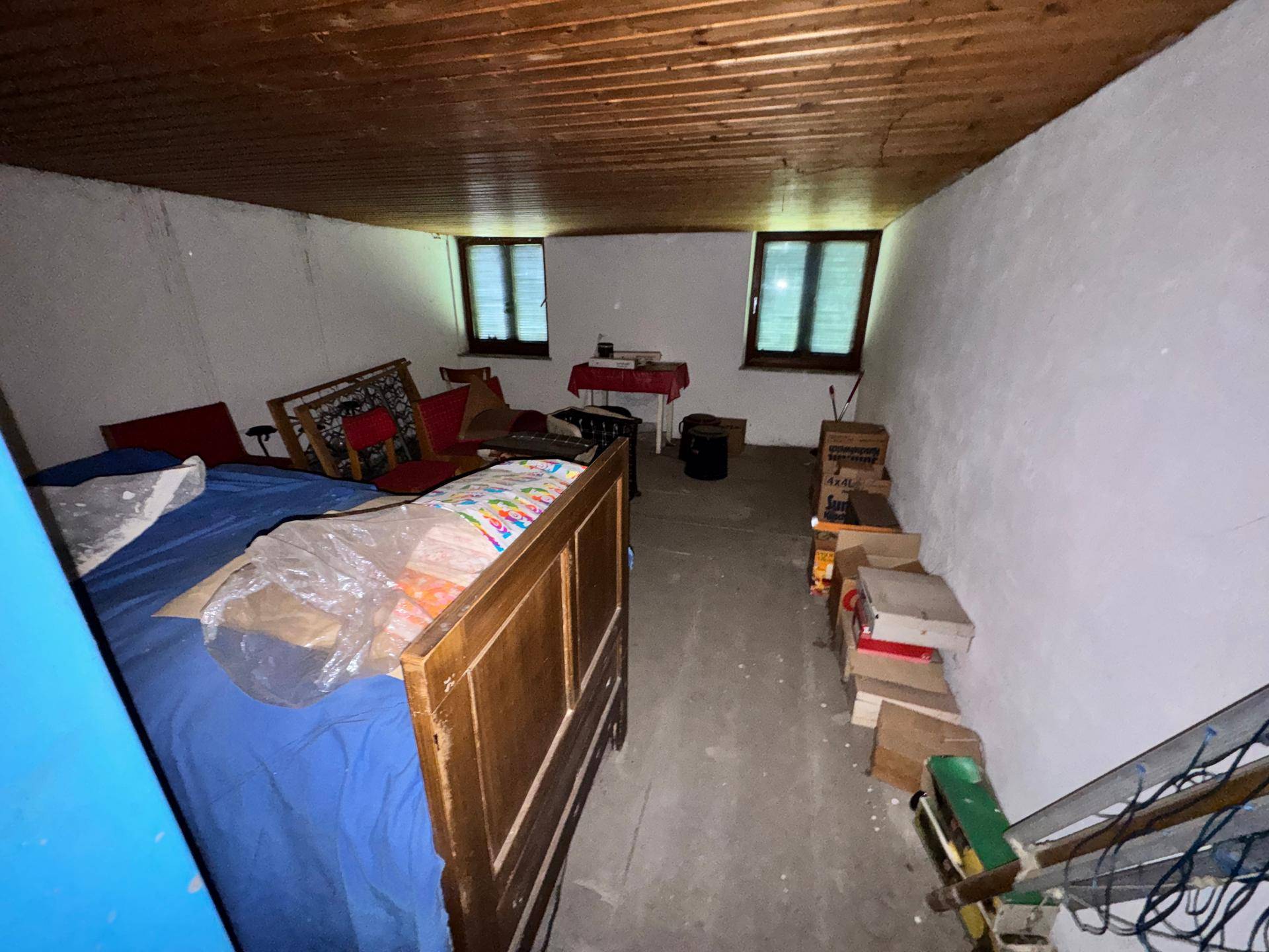 Einfaches Schlafzimmer mit Holzbalkendecke, Betonboden und zwei kleinen Fenstern in rustikalem Zustand.
