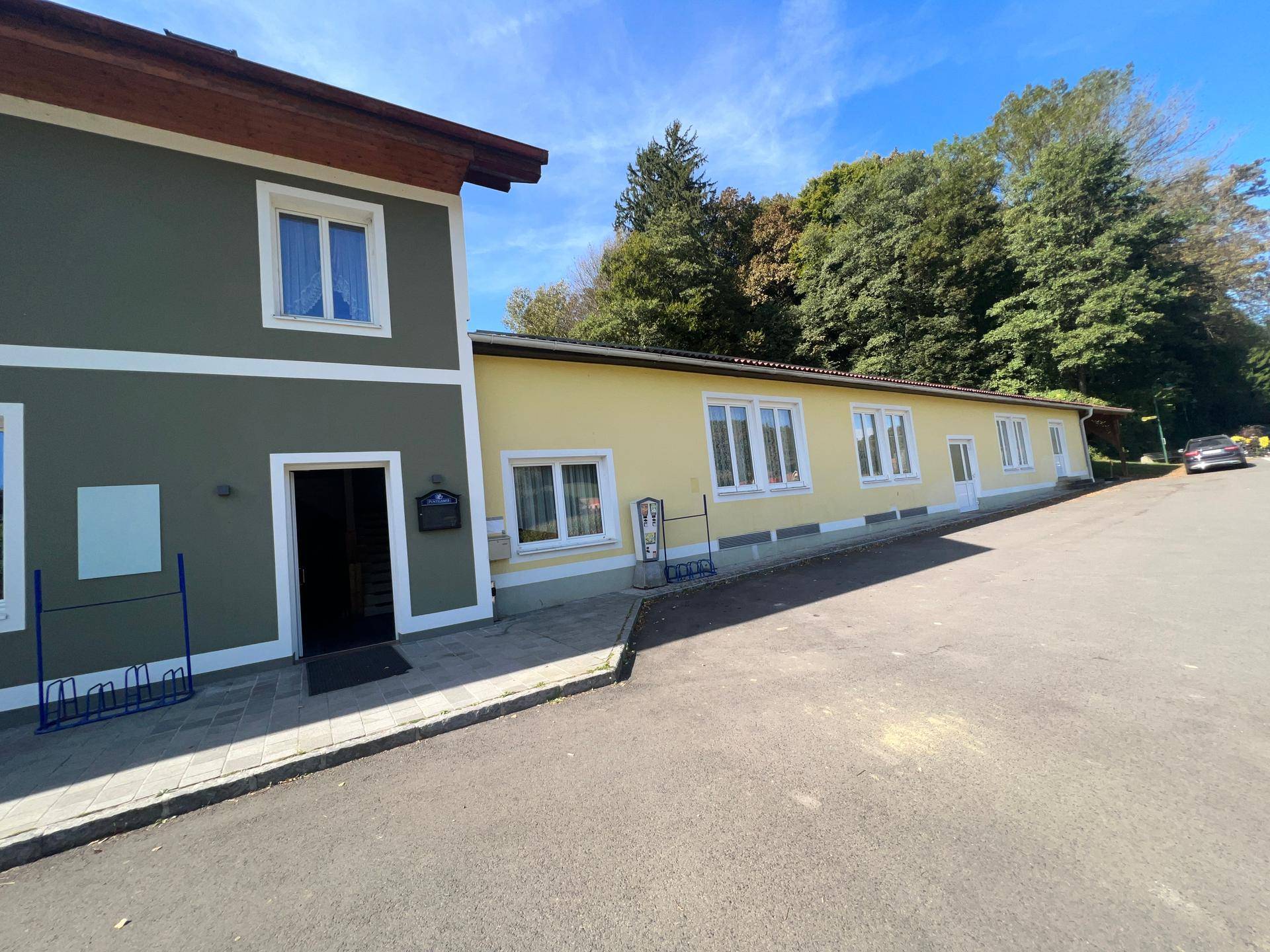 Seitenansicht des Gebäudes mit gelber Erweiterung und Eingangsbereich, umgeben von Bäumen.