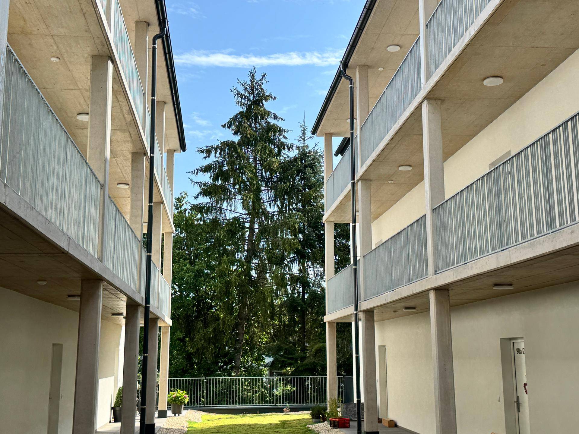 Blick zwischen zwei Gebäudeteilen auf hohe Bäume und eine gepflegte Rasenfläche.