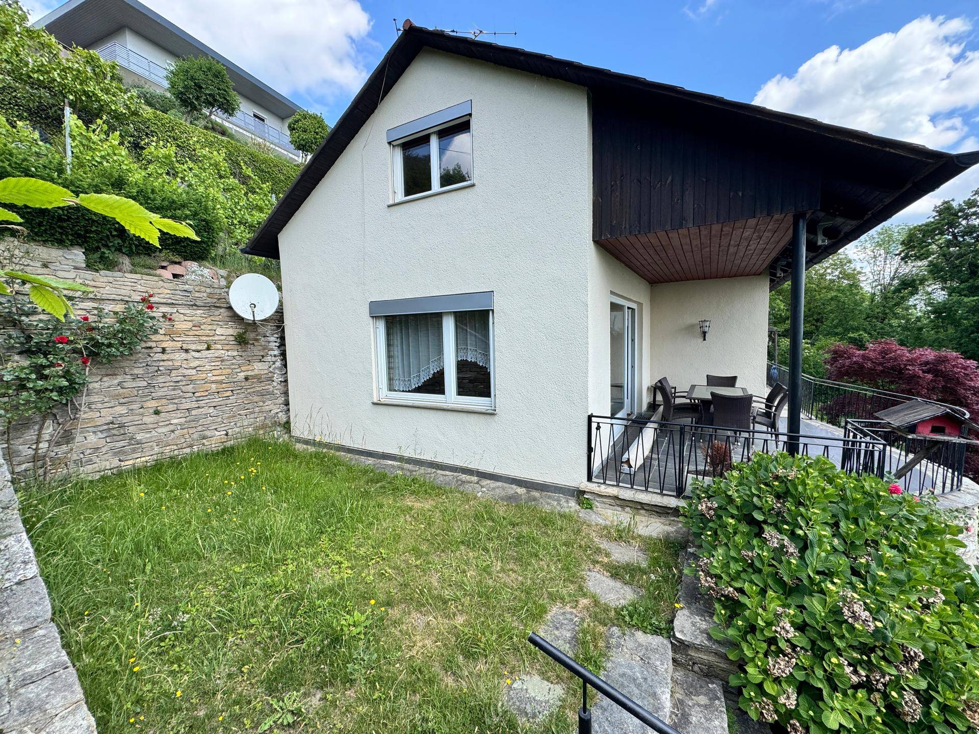 Ansicht des Hauses von unten mit kleiner Rasenfläche und Balkon, umgeben von grünen Büschen.