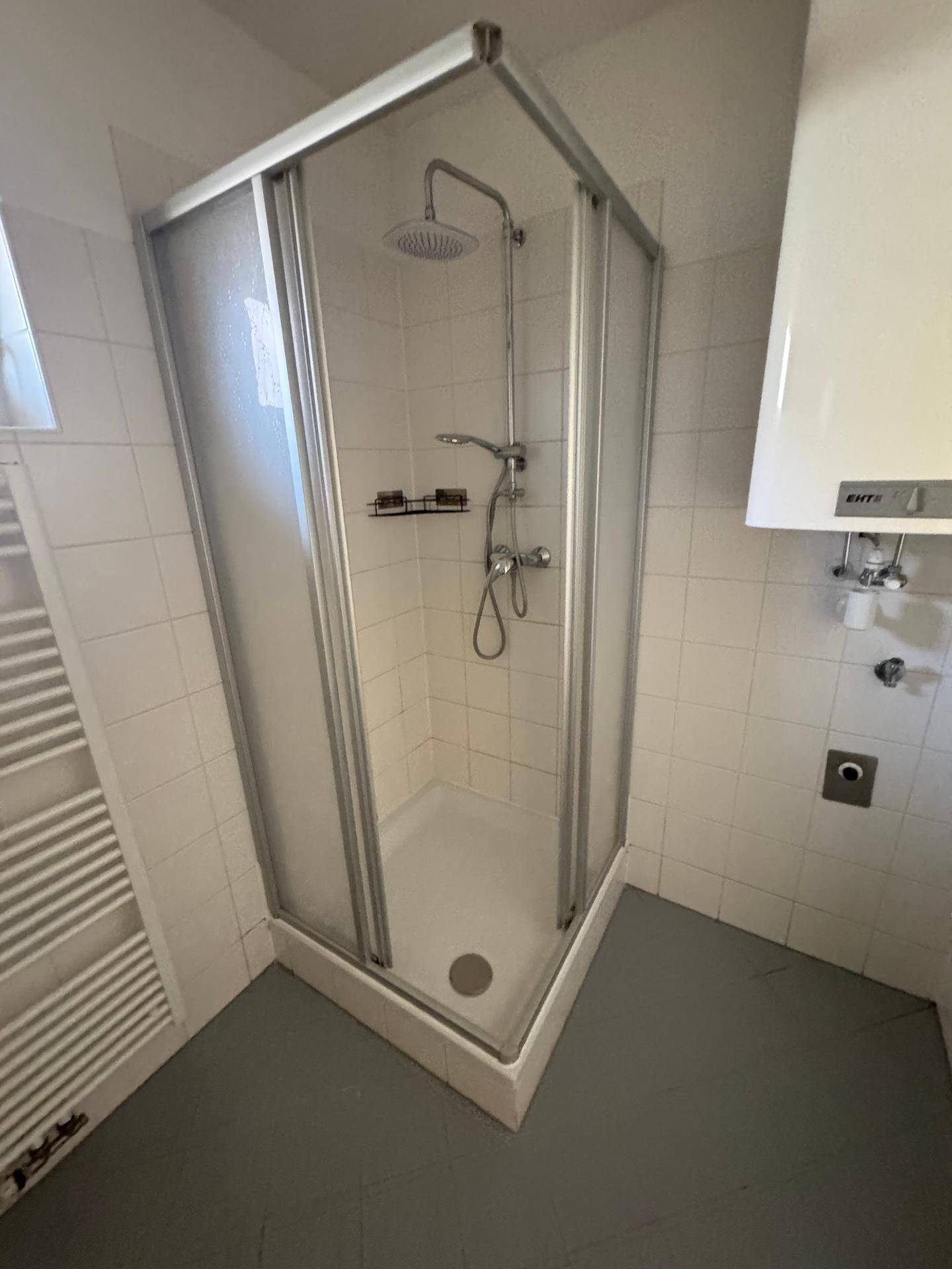 Badezimmer mit Eckduschkabine, weißen Fliesen, Handtuchheizkörper und Warmwasserbereiter.