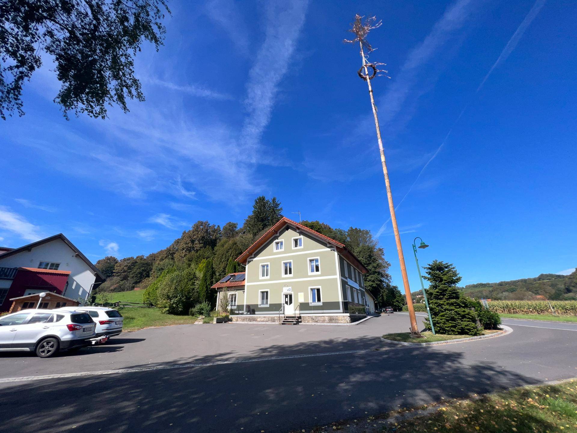 Weitläufige Außenansicht des Gebäudes mit Parkplätzen und einem Maibaum unter blauem Himmel.
