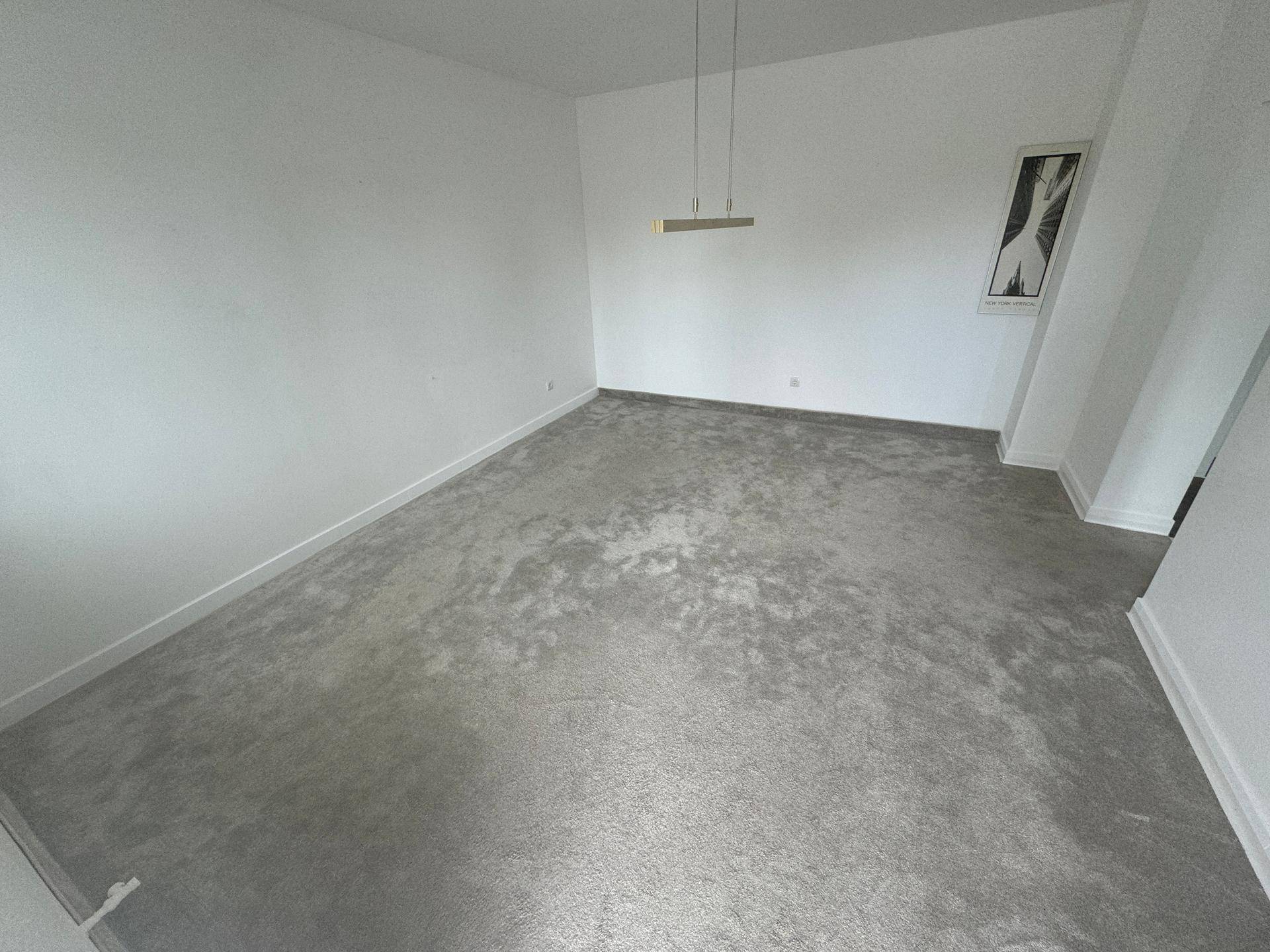 Heller Raum mit grauem Teppichboden, weißen Wänden und einer modernen Hängelampe als Blickfang.