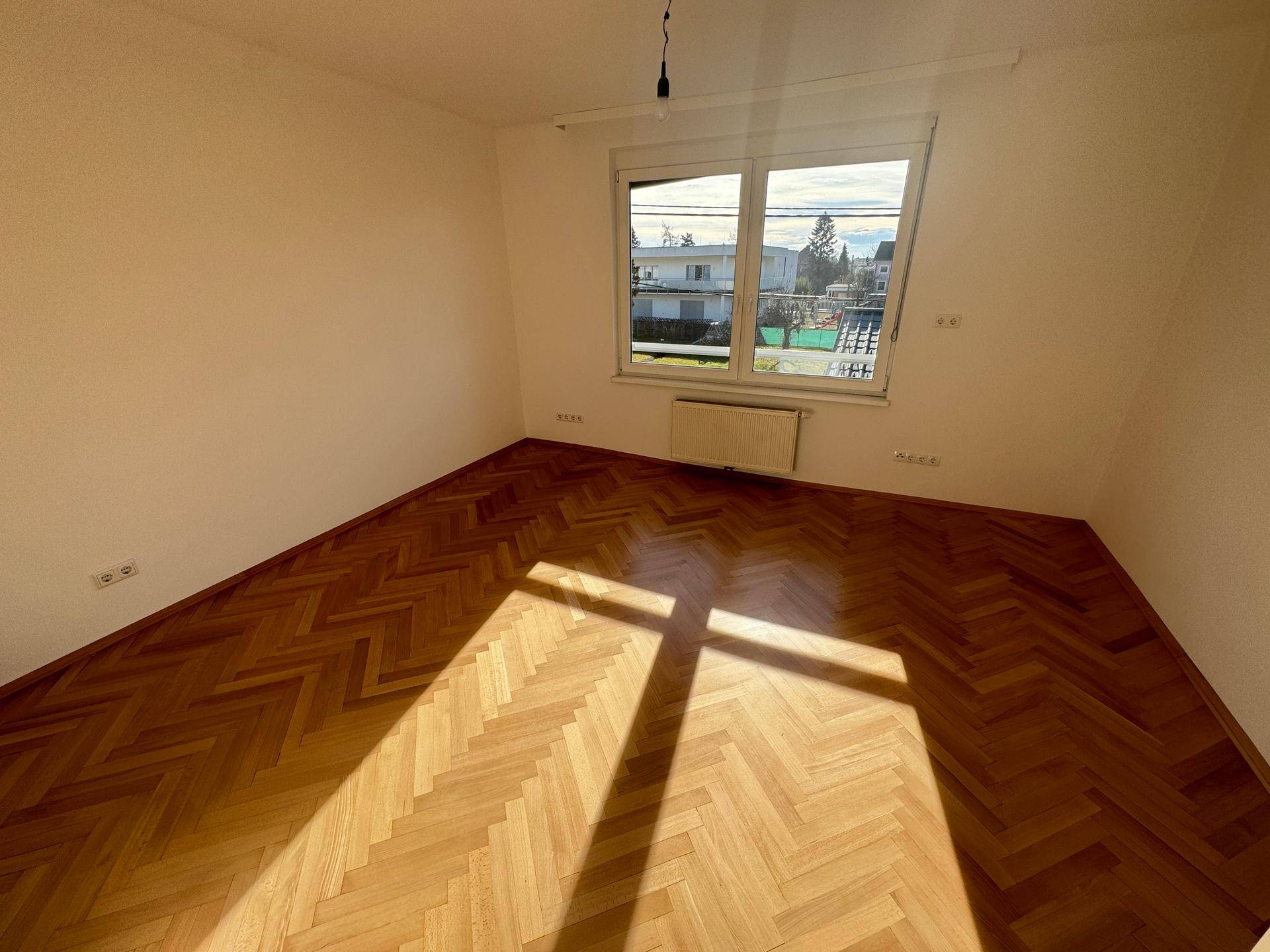 Heller Raum mit Parkettboden, großem Fenster und Sonneneinstrahlung.