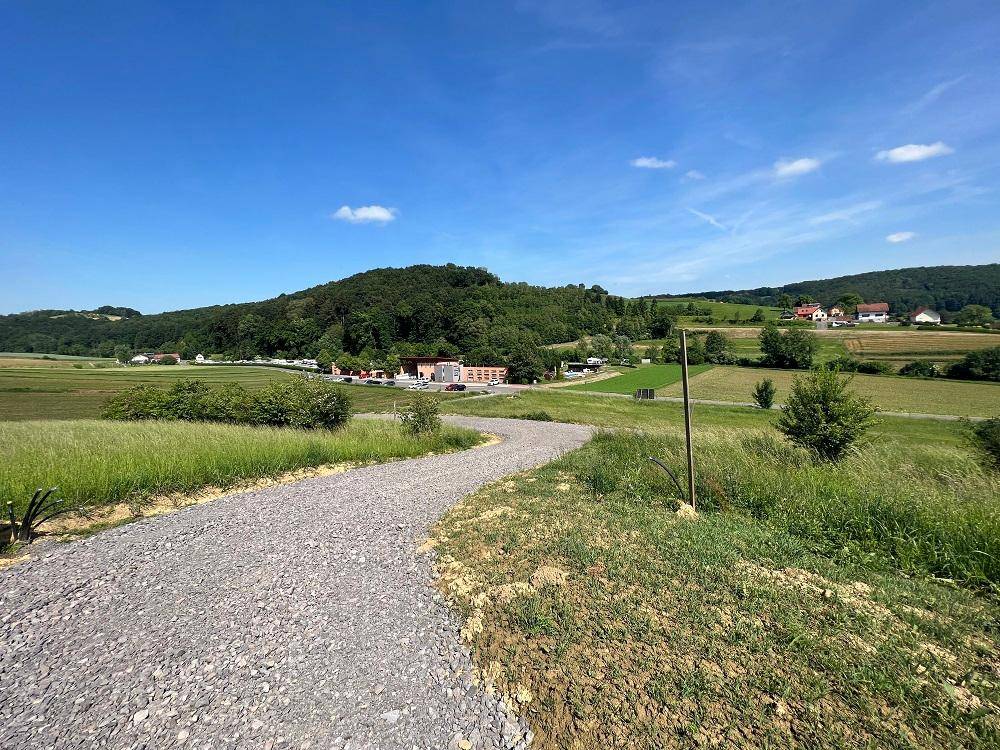 Geschwungener Schotterweg führt durch eine grüne Landschaft mit Häusern in der Ferne.