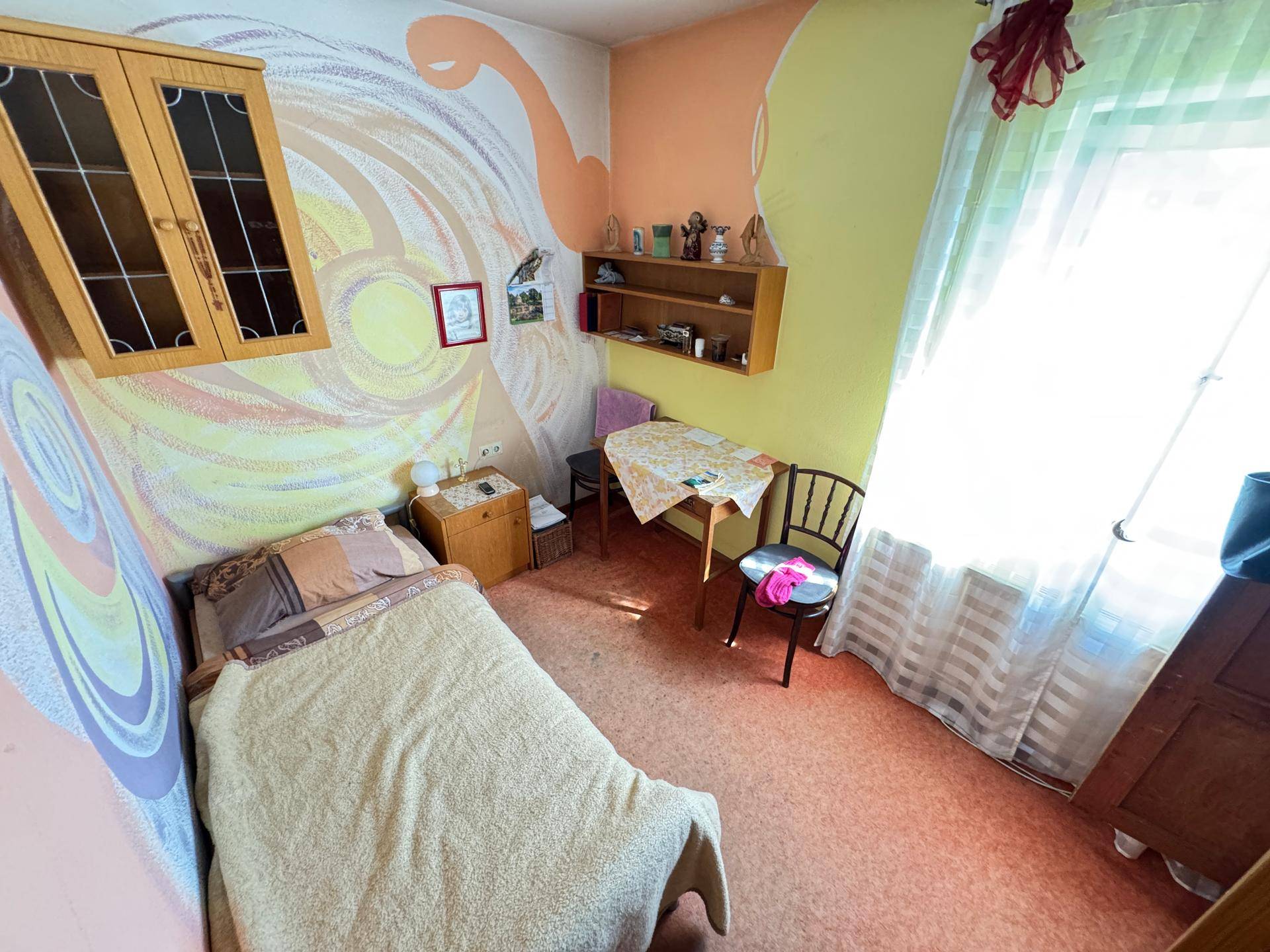 Kinderzimmer mit farbenfroher Wandgestaltung, Einzelbett, kleinem Tisch und einem Fenster.