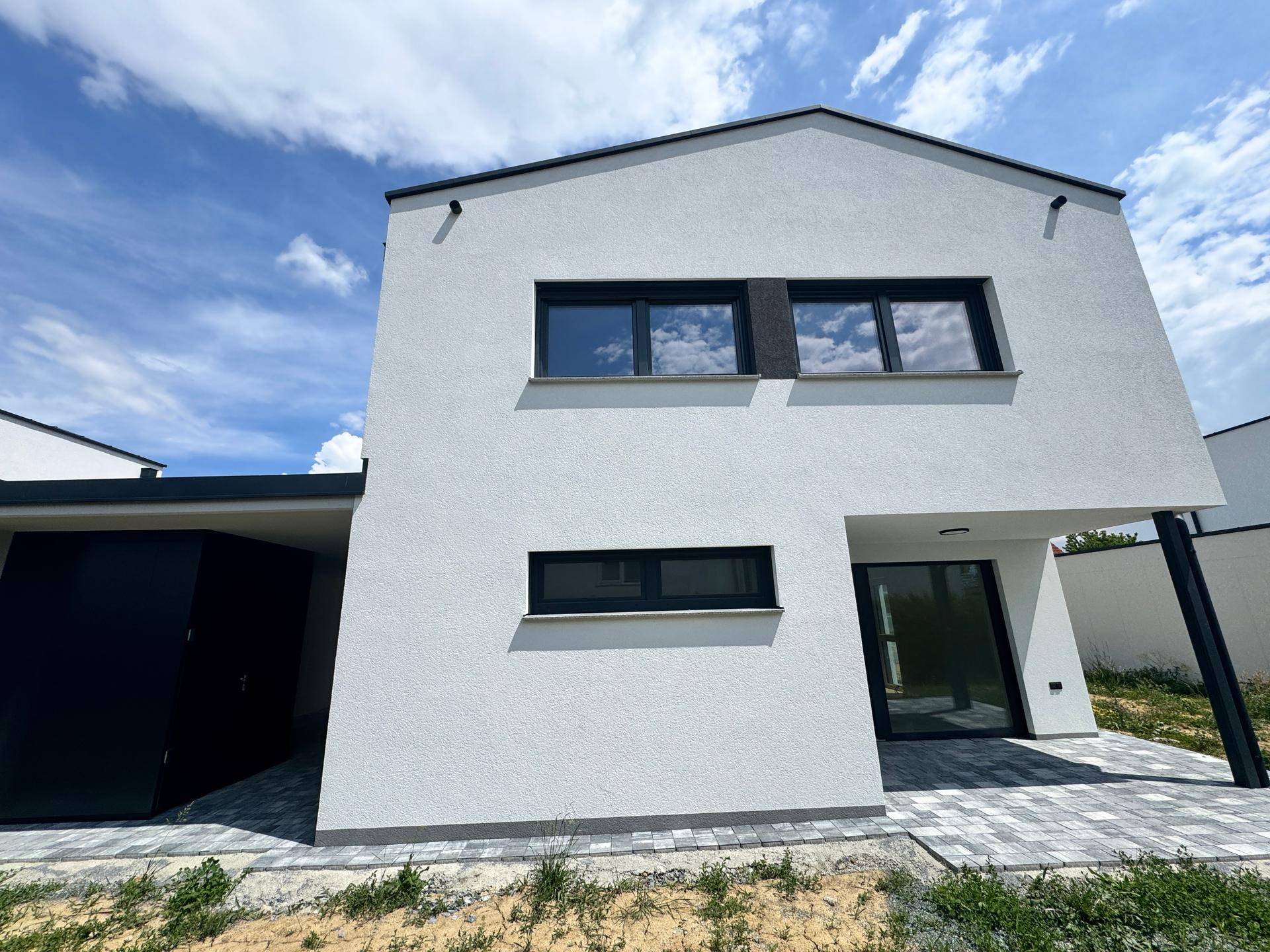 Ansicht der modernen Hausfassade mit großen Fenstern und gepflastertem Bereich vor dem Eingang.
