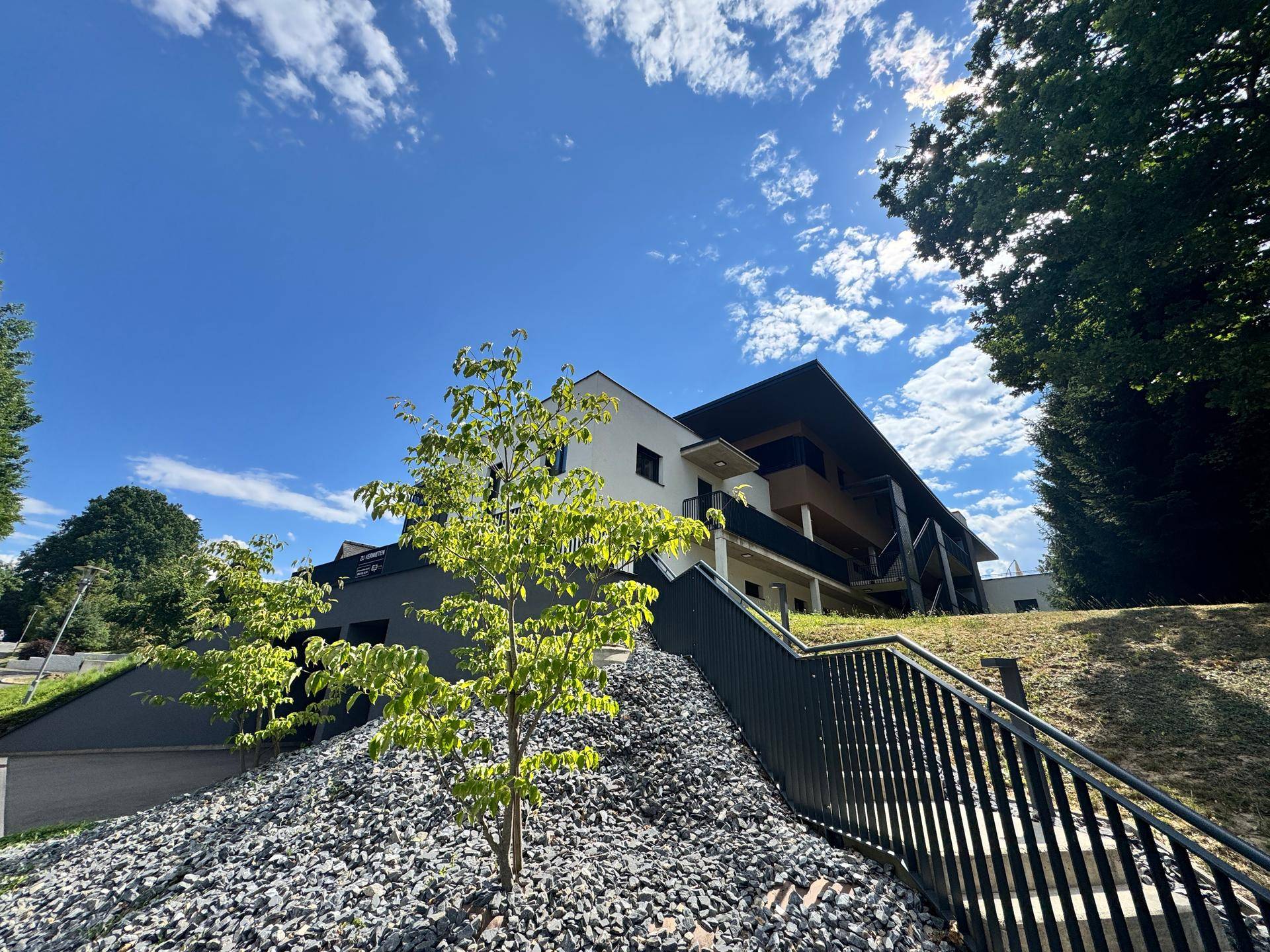 Neubau-Gebäude mit moderner Architektur, dunklen Balkonen und einer begrünten Hanglage unter blauem Himmel.