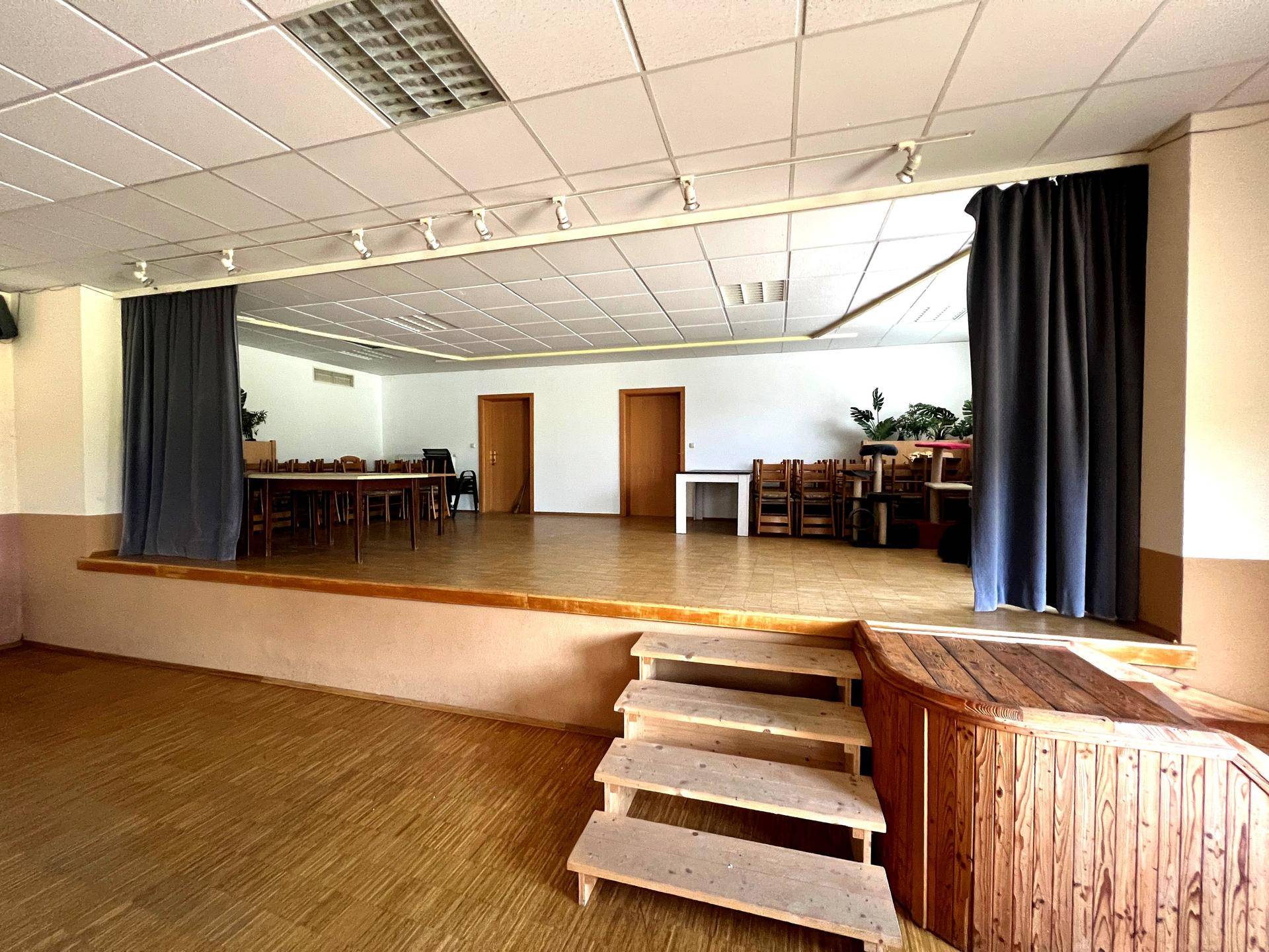 Ein großer Veranstaltungssaal mit Bühne, Holzboden und Vorhängen, ideal für Aufführungen oder Versammlungen.