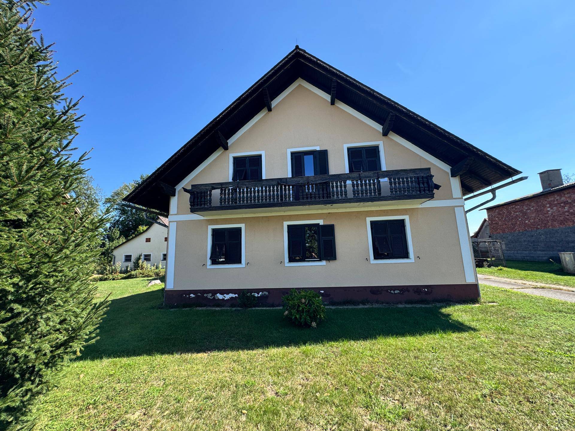 Haus mit Balkon, grüner Wiese und kleinem Nadelbaum im Vorgarten unter blauem Himmel.