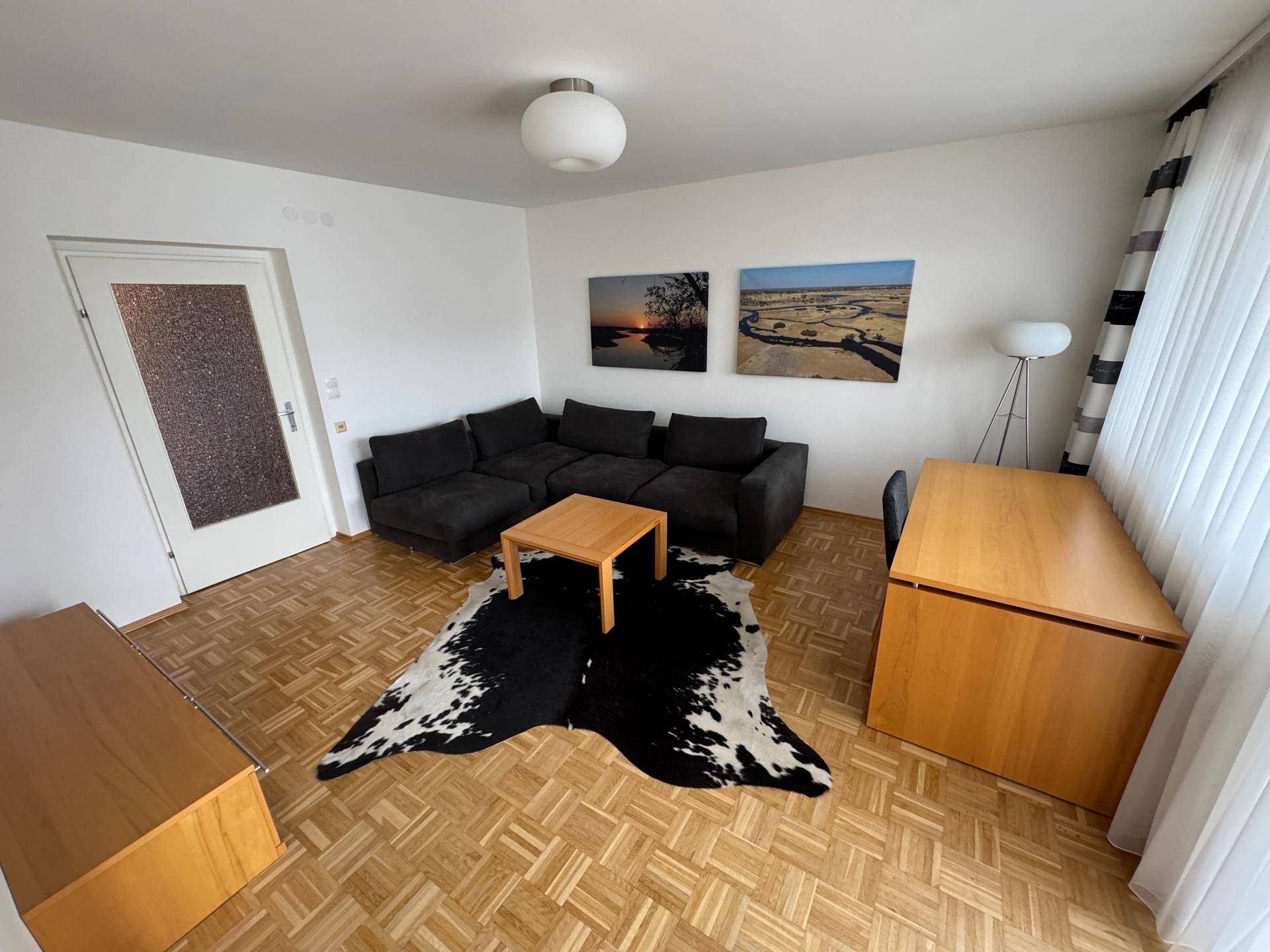 Helles Wohnzimmer mit dunkler Eckcouch, Holztisch und Arbeitsbereich am Fenster.