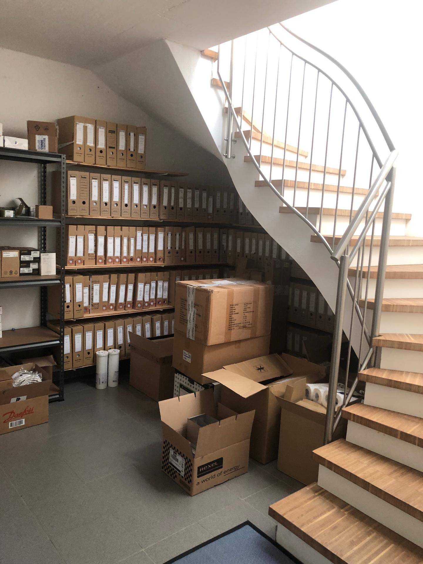 Lagerbereich unter der Treppe mit Regalen voller Aktenordner und Kartons.