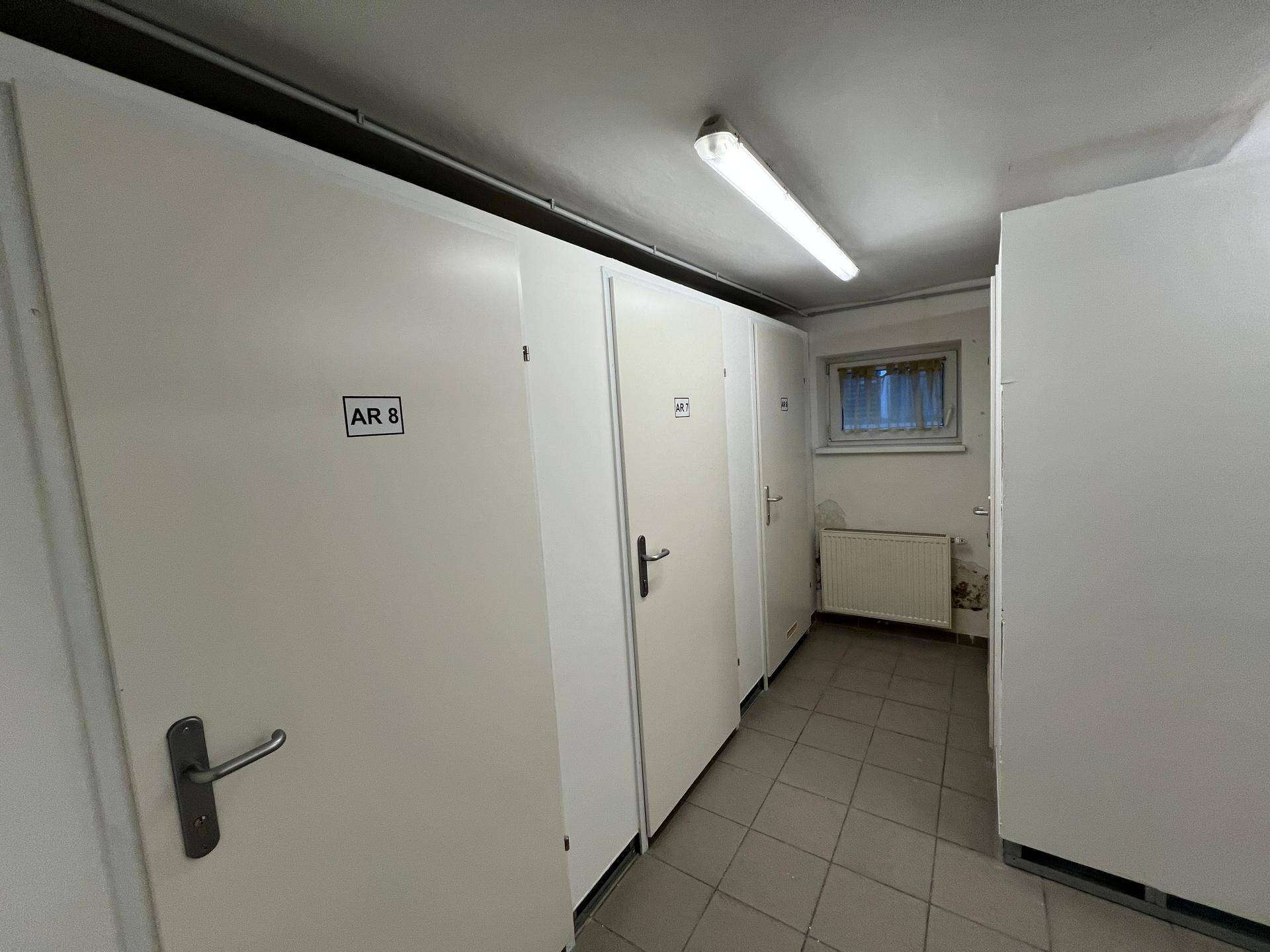 Langer Kellerflur mit vielen weißen Lagerabteilen, Fliesenboden, Fenster und Heizkörper.