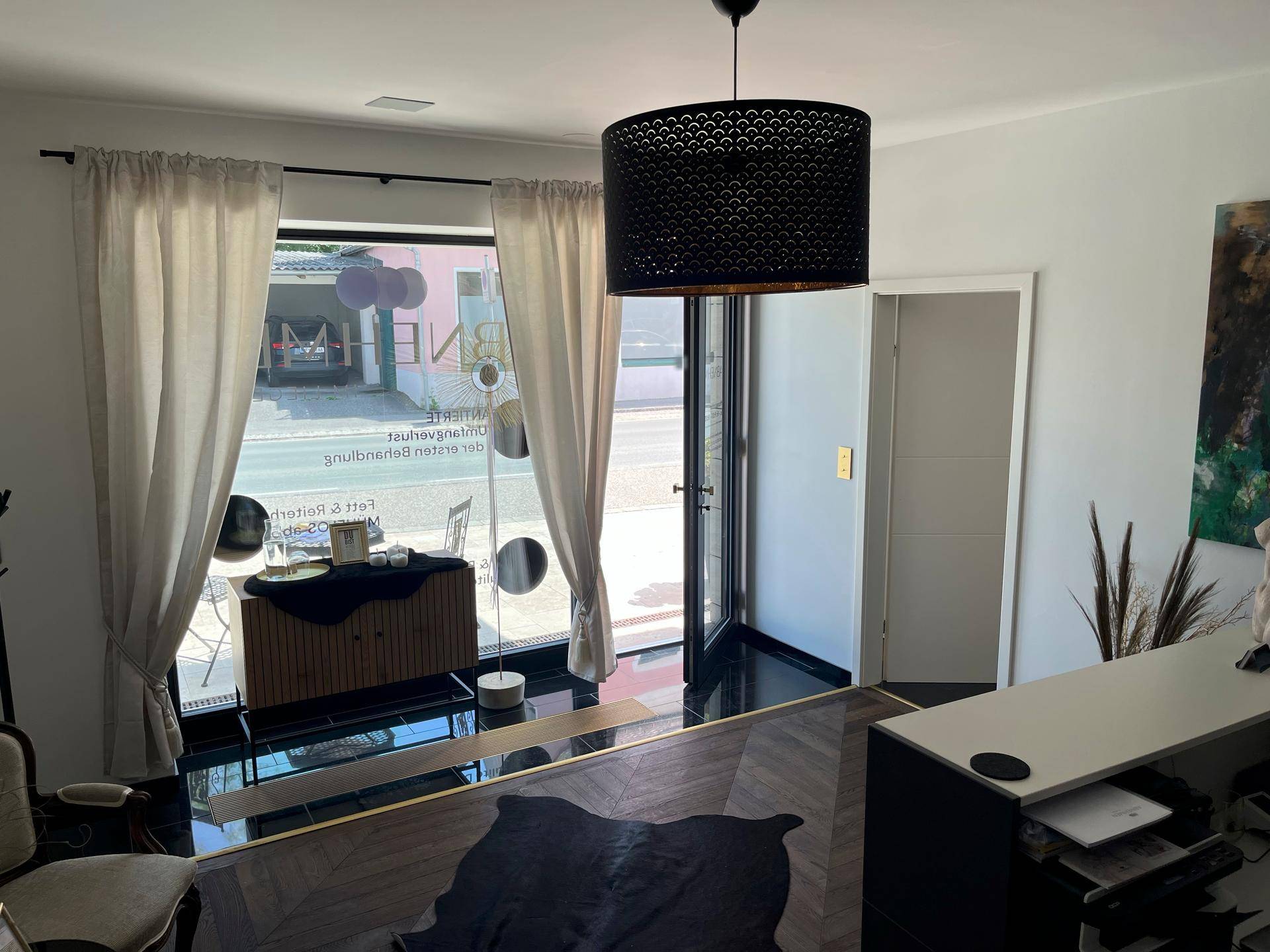 Empfangsbereich mit dunklem Boden, hellen Vorhängen und Blick auf die Straße durch große Fenster.
