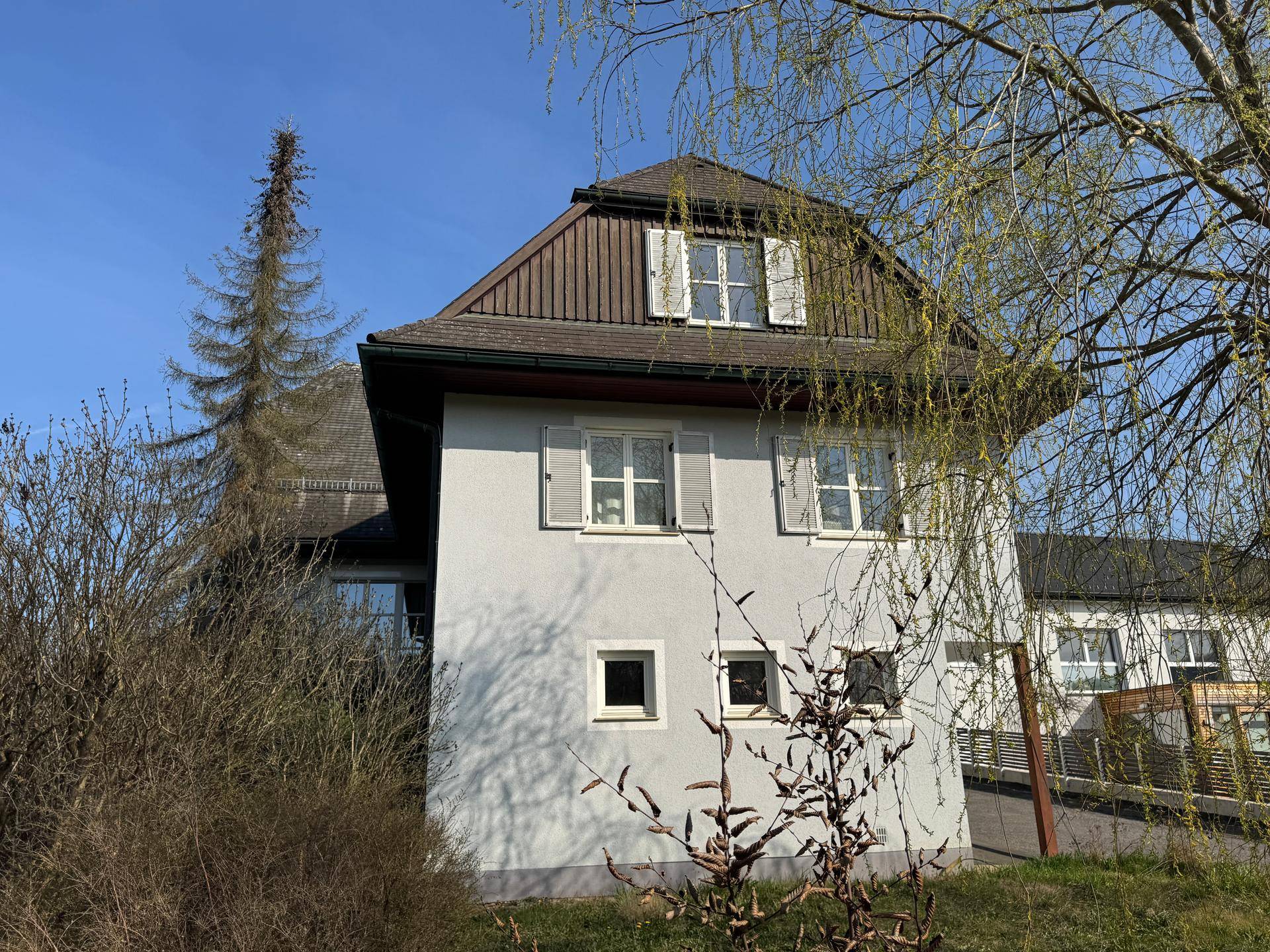 Haus mit heller Fassade, Mansarddach und grüner Umgebung unter blauem Himmel.