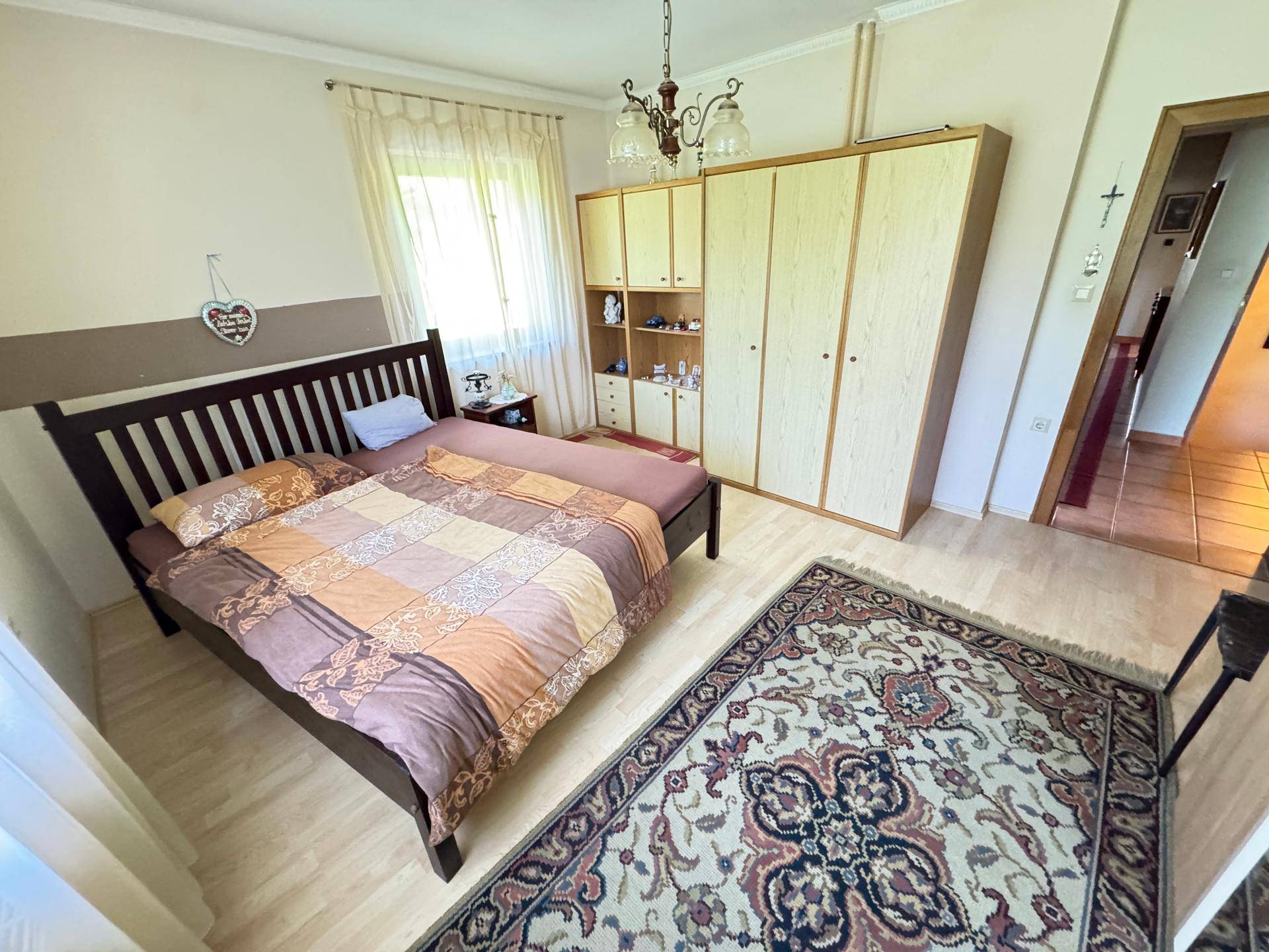 Schlafzimmer mit Doppelbett, Teppich, Holzschrank und einem Fenster, das viel Tageslicht hereinlässt.