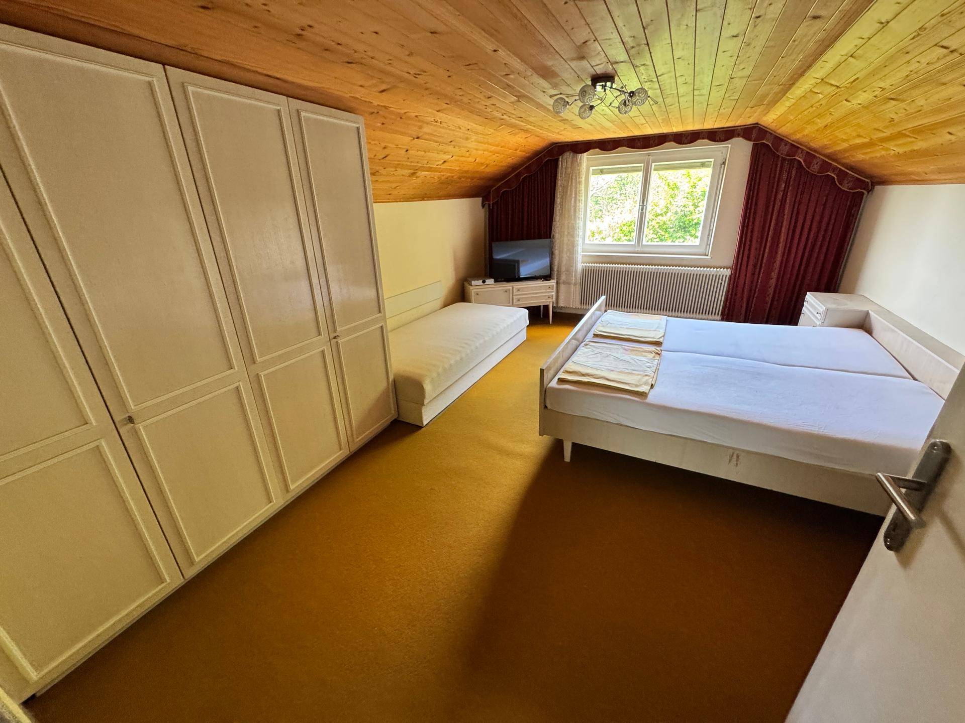 Schlafzimmer mit Holzdecke, Teppichboden und zwei Betten unter einem Fenster.