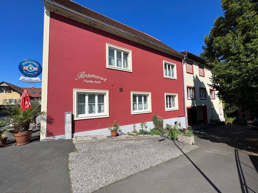 Rotes Gebäude mit weißen Fenstern und einem Schild "Rosenwirt" an einer kleinen Zufahrt.